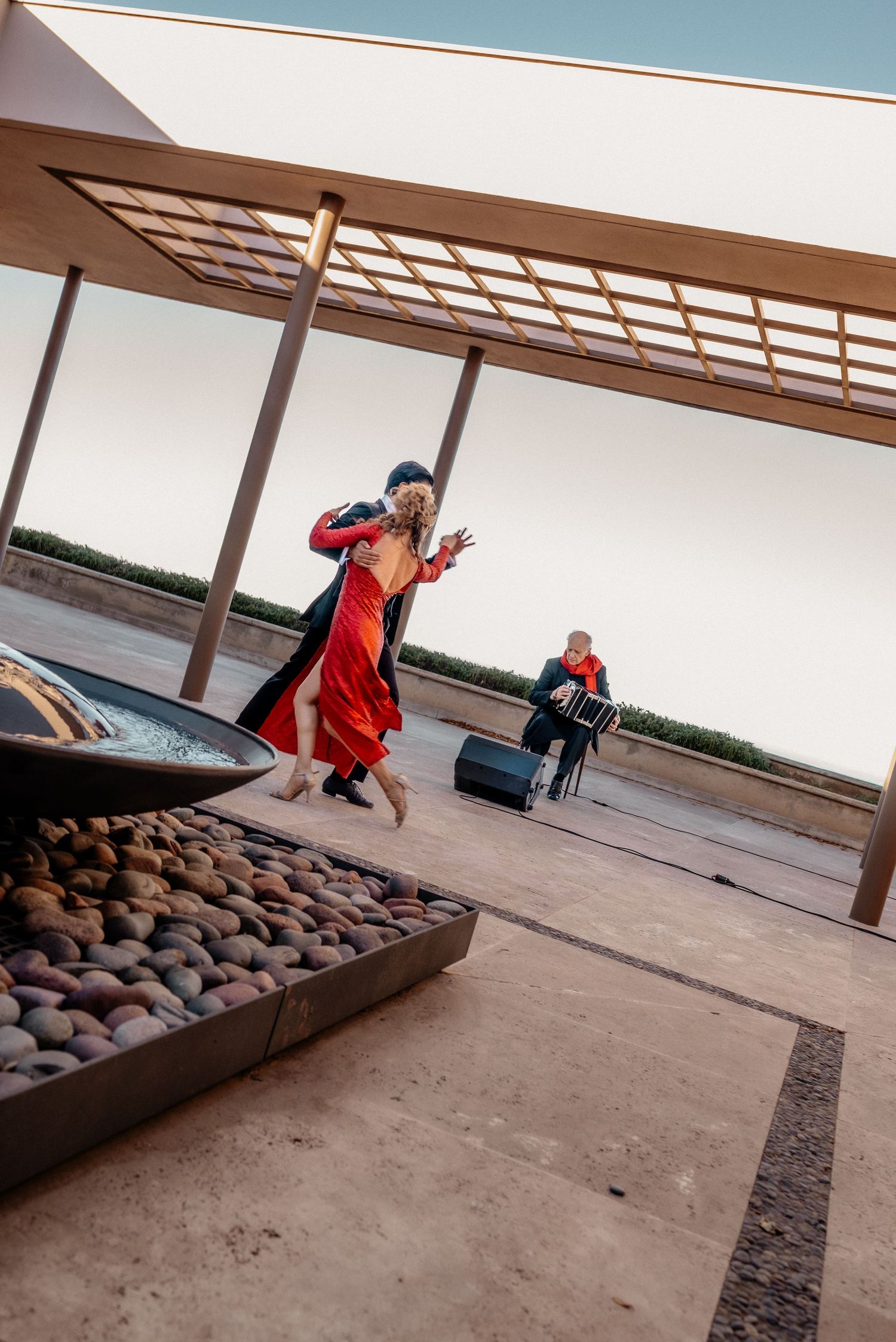 Tango dancers in red and black perform outdoors under a wooden pergola while a musician plays accordion in the background.