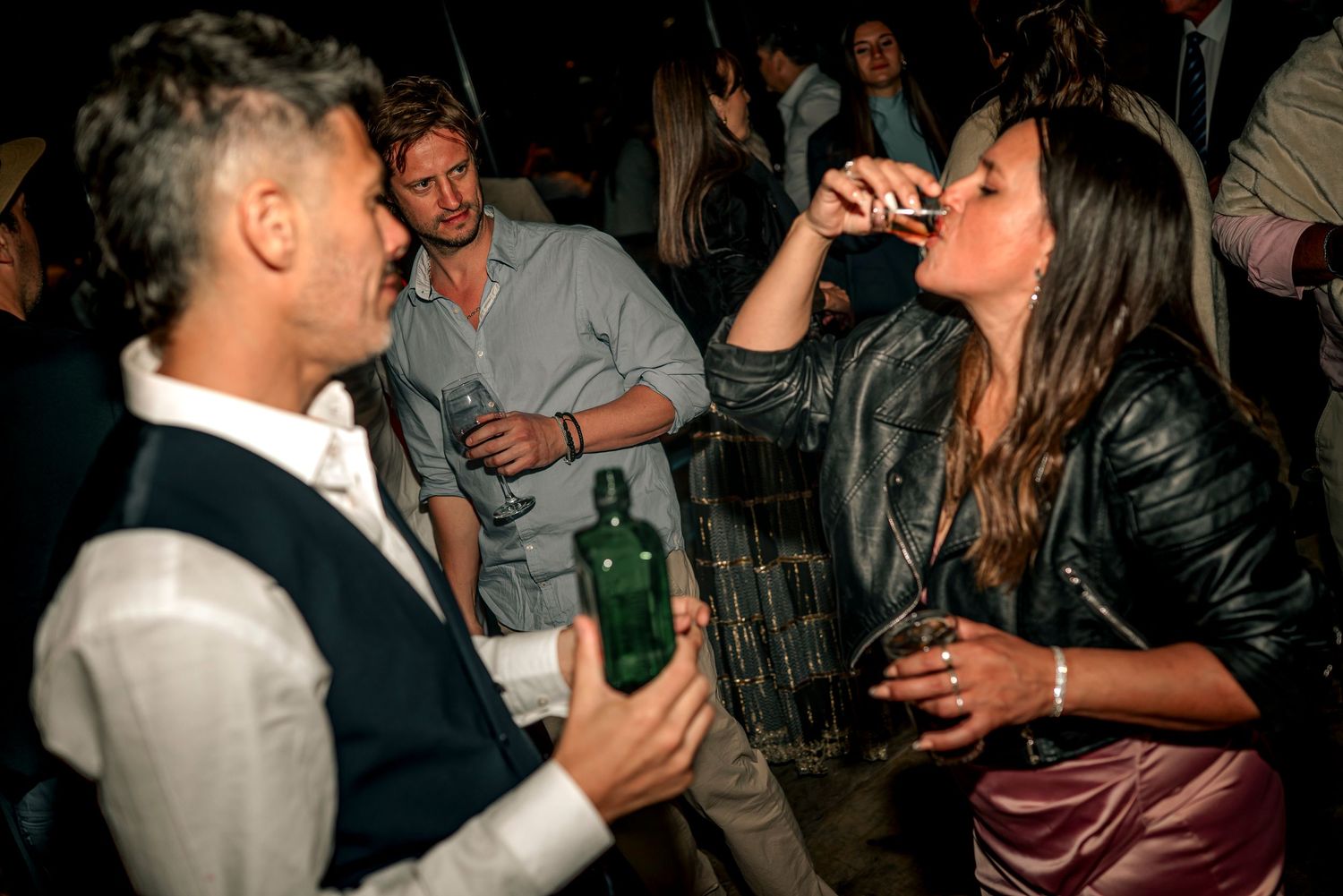 People drinking and socializing at a crowded nightclub or bar event with bottles and drinks visible.