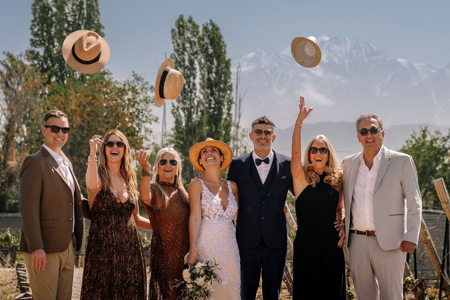 Wedding party members pose outdoors in formal attire with mountains and trees visible in the background.