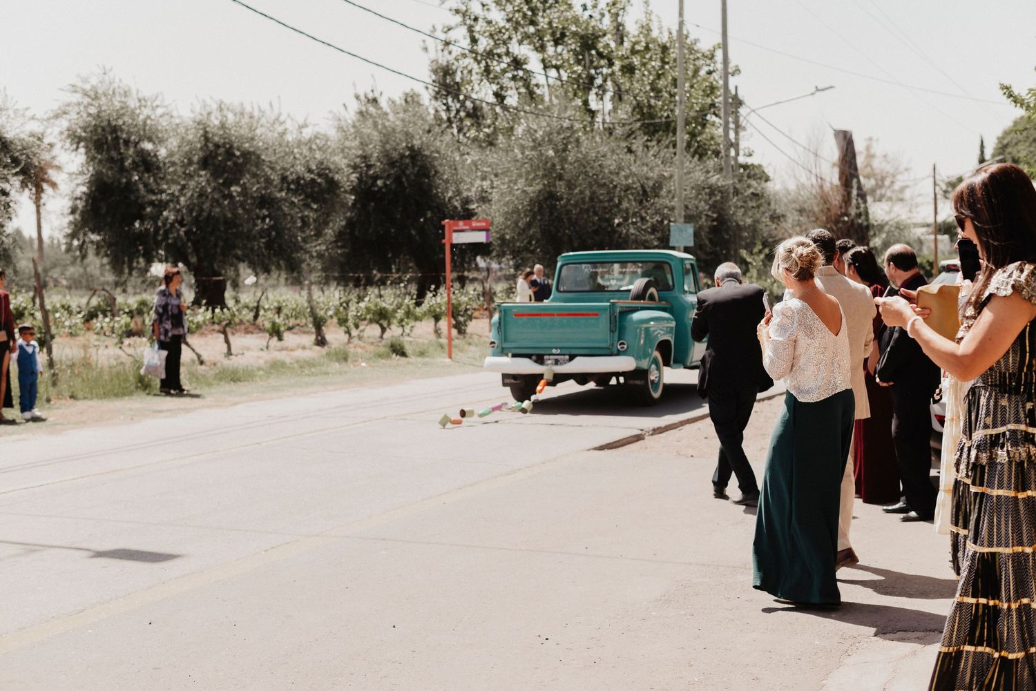 A vintage turquoise car is parked on a sunny street with trees and people gathered in the background.