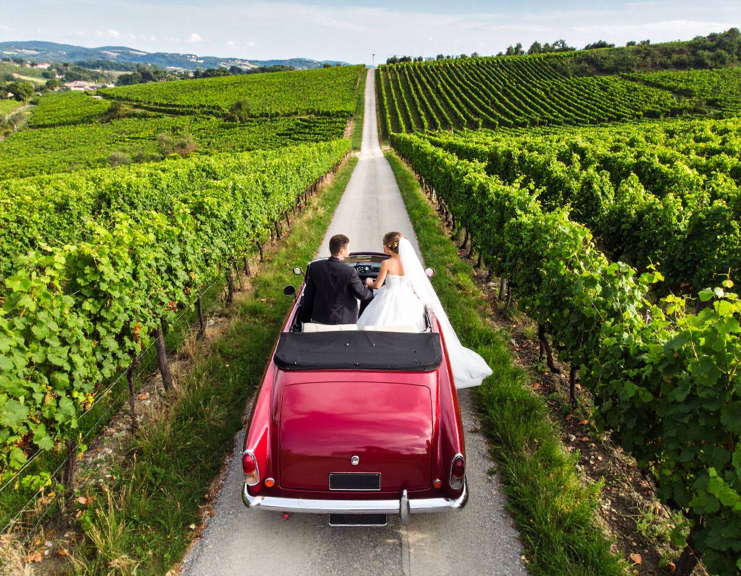 Vintage red convertible car driving through scenic vineyard rows and green rolling hills of wine country.