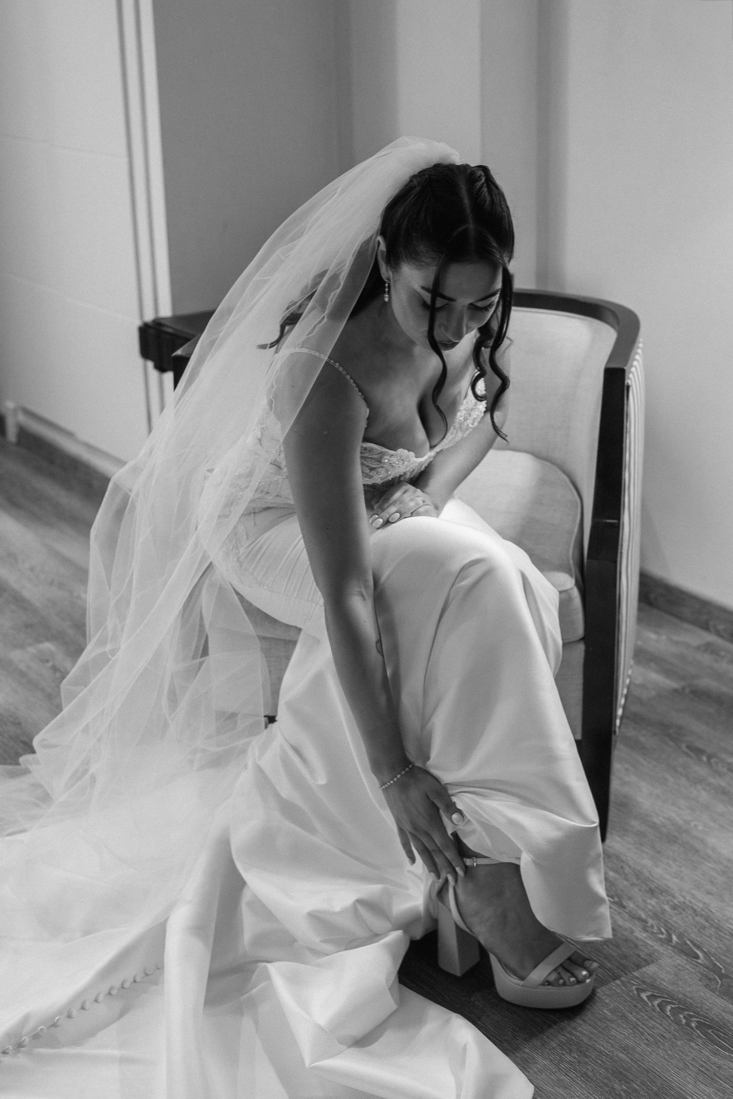 La novia, vestida con un traje blanco y velo, está sentada en una silla abrochándose el zapato de novia; la imagen, capturada en elegante