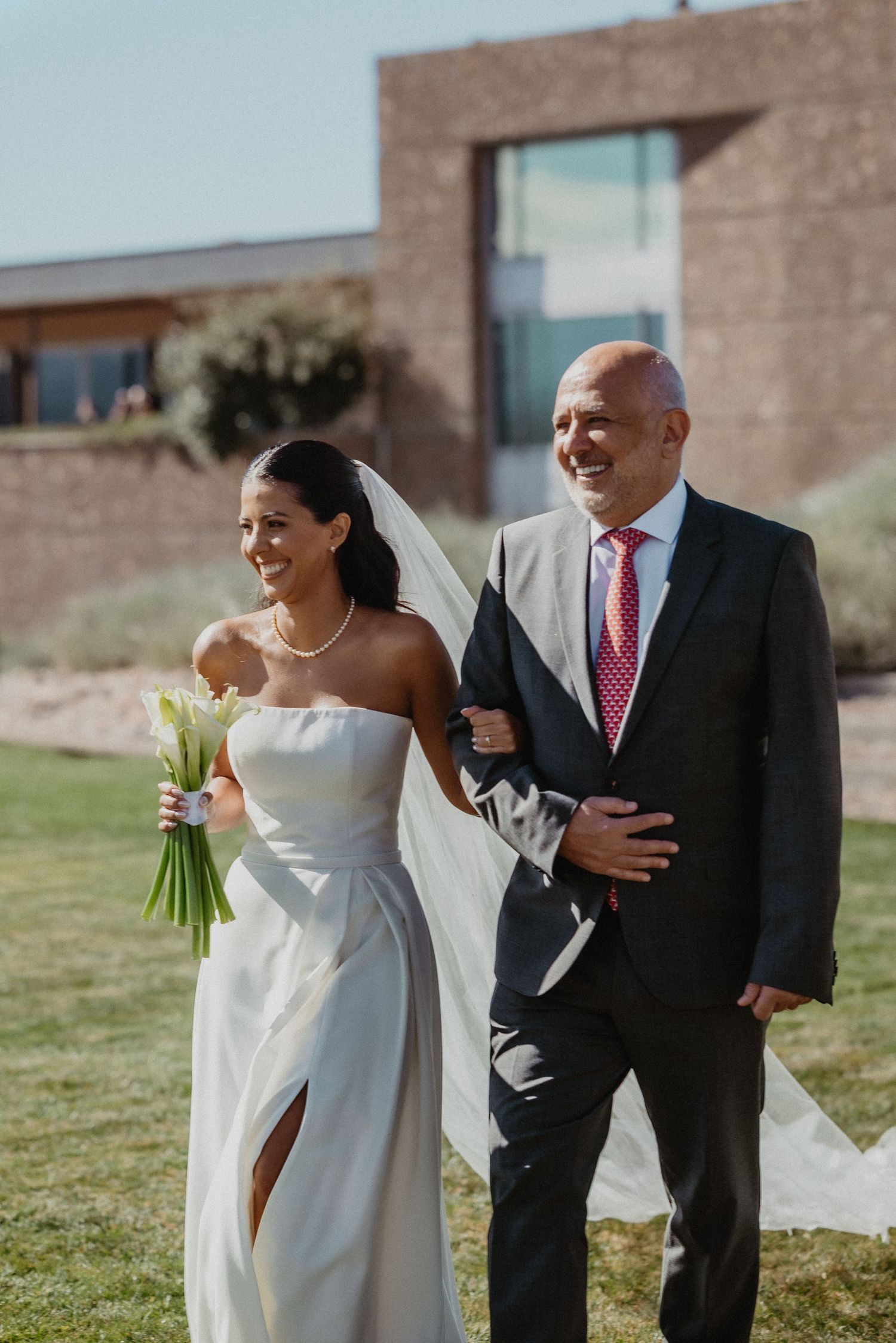 Bride in white strapless gown with veil and bouquet walks arm-in-arm with suited man on grass at outdoor wedding.