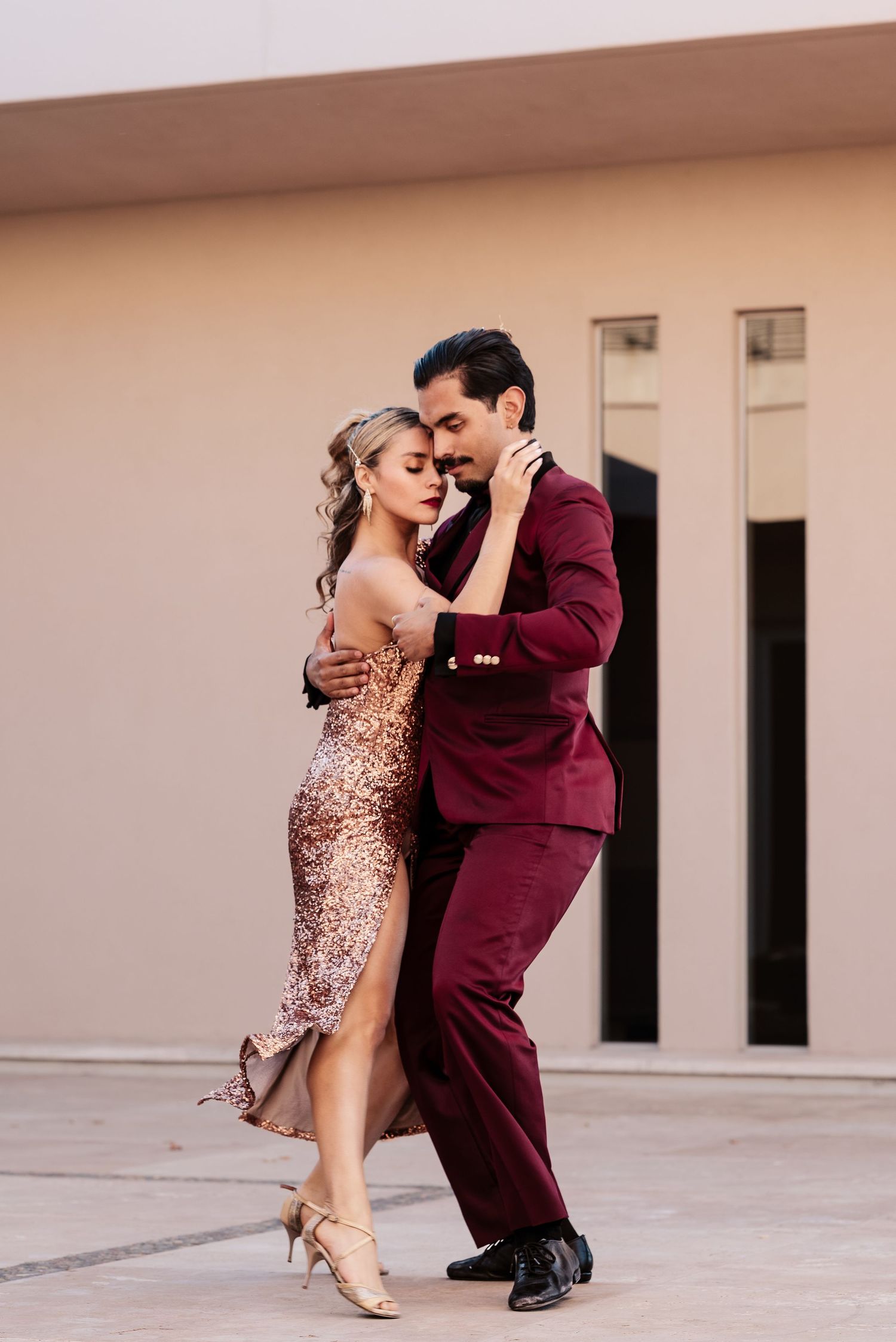 Elegant couple dancing tango outdoors, woman in gold sequin dress, man in burgundy suit, romantic pose.