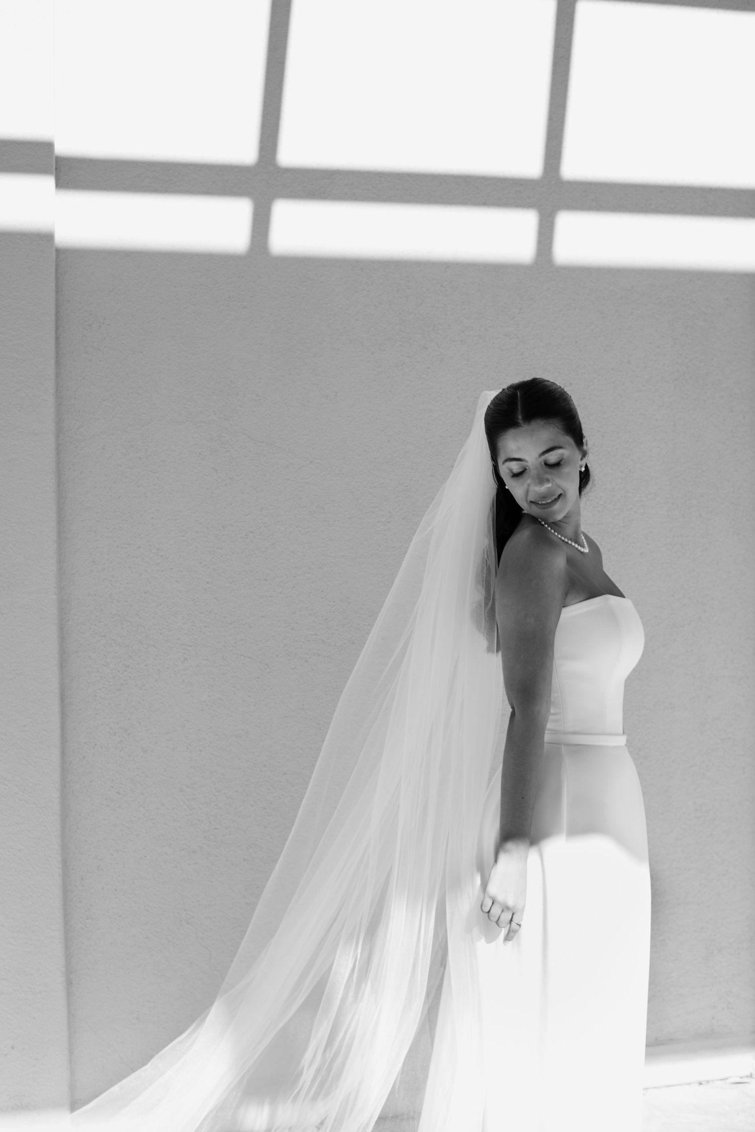 Black and white photo of a bride in a strapless gown with a long flowing veil, smiling softly in elegant natural light.