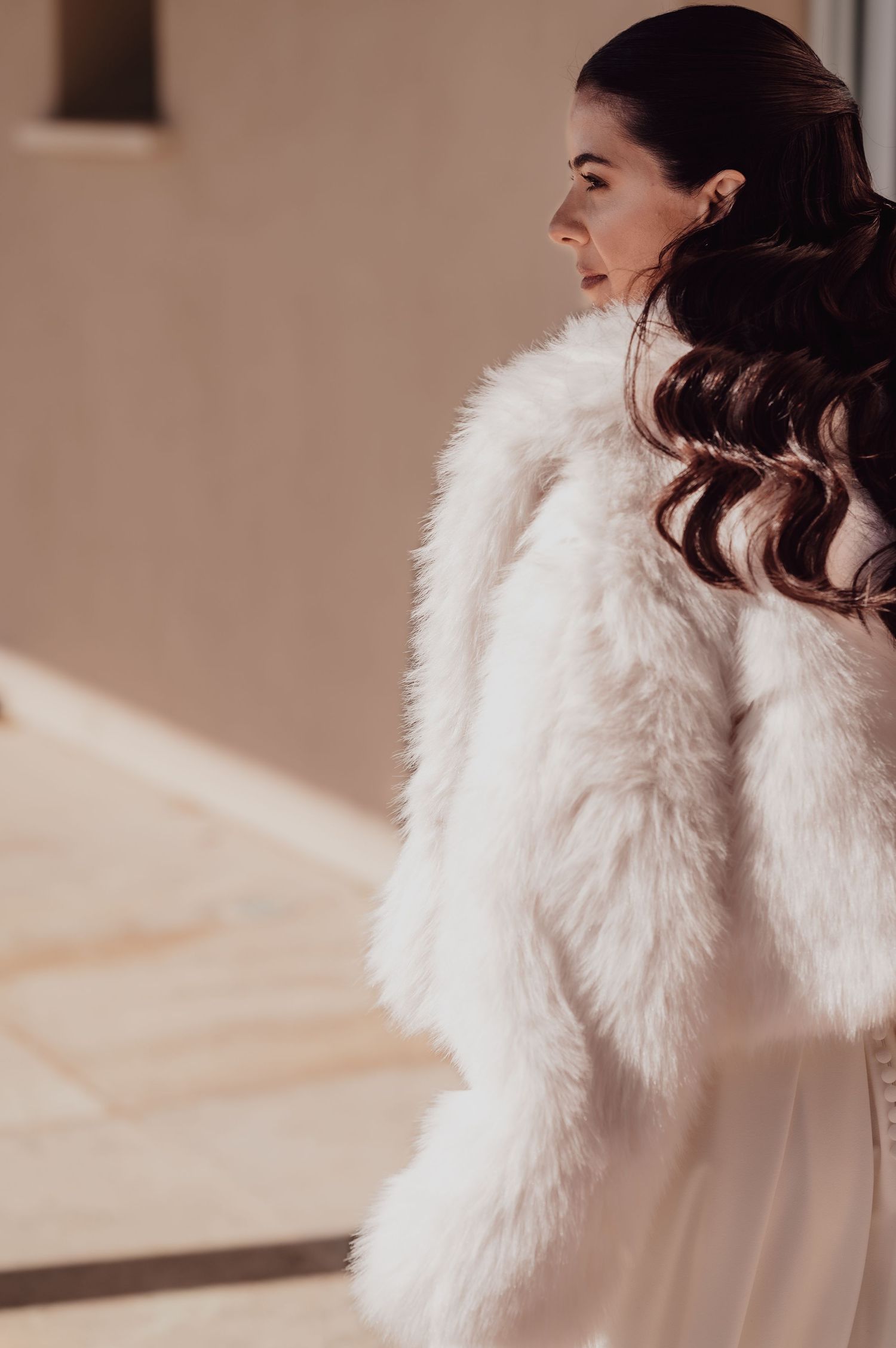 Bride in white faux fur coat with dark curly hair, elegant bridal look in warm natural light.