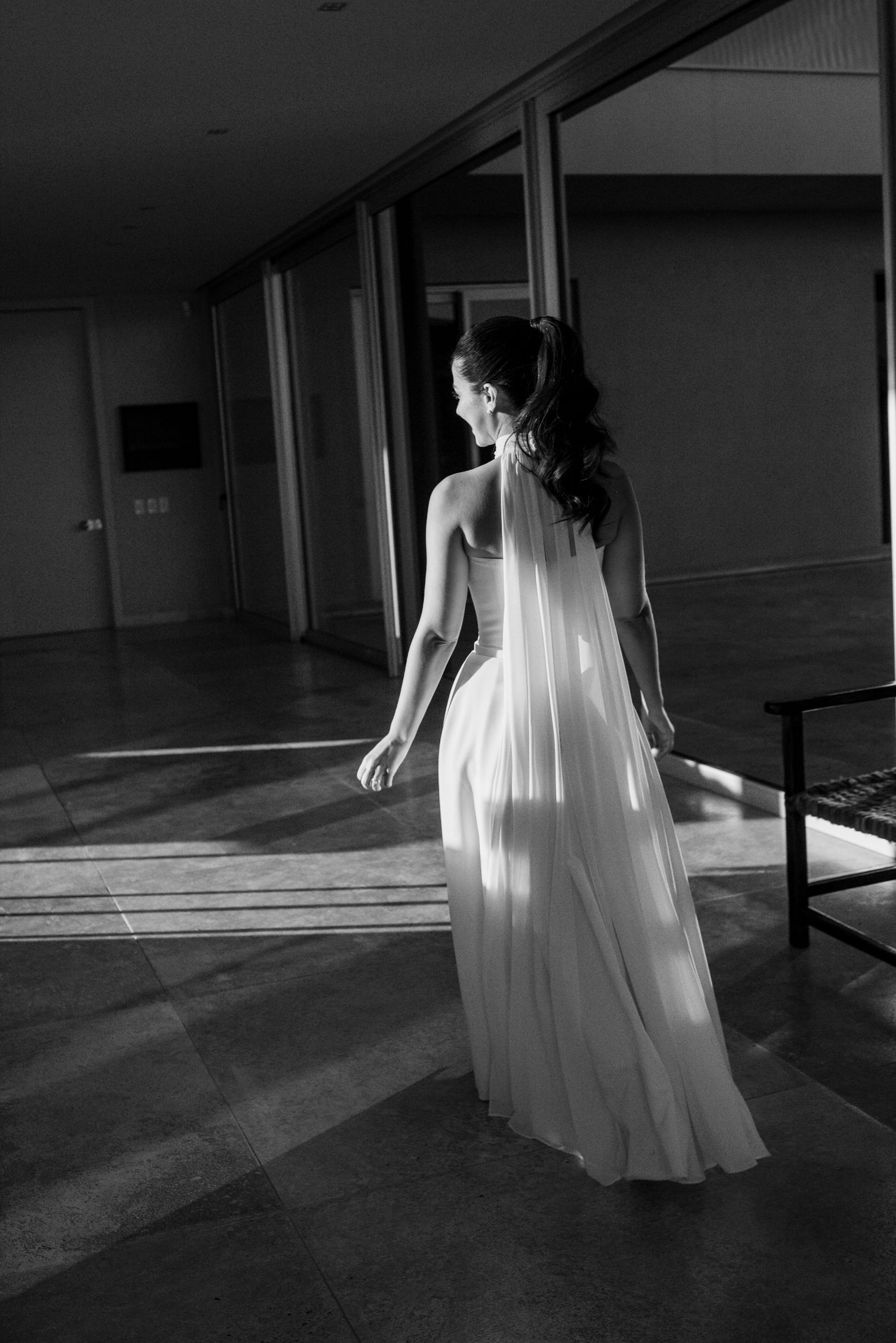 Black and white photo of a bride in a flowing white gown standing in a sunlit hallway, viewed from behind.