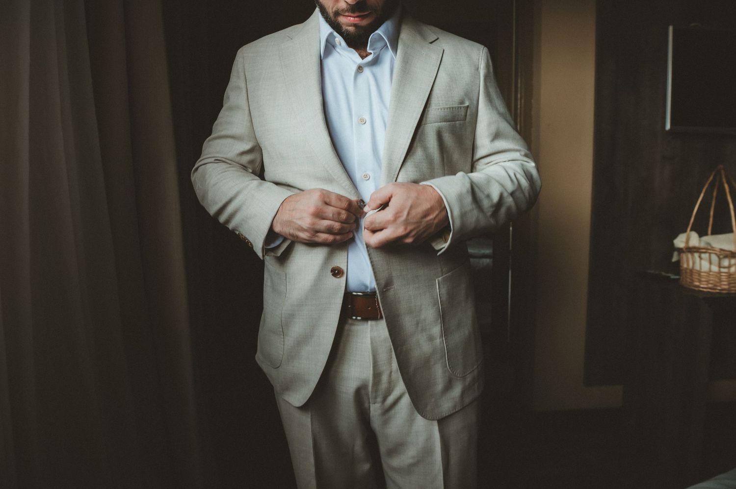 A person wearing a light gray suit and white shirt adjusts their cuffs while standing in a dimly lit room.
