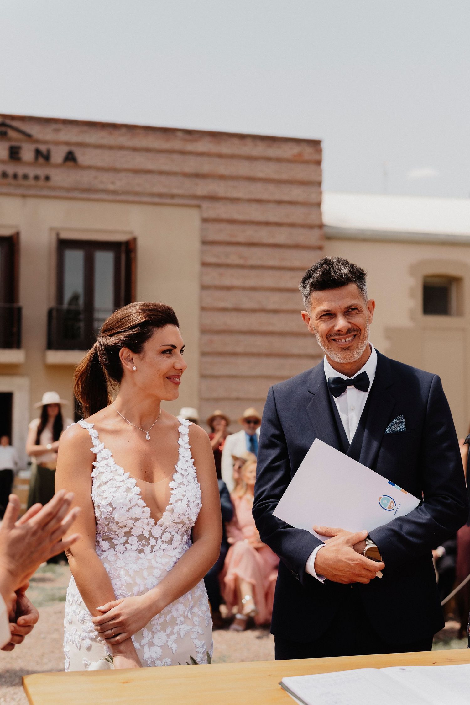 A couple at their wedding ceremony standing at an outdoor venue, with the bride in a lace dress and groom in a bowtie.