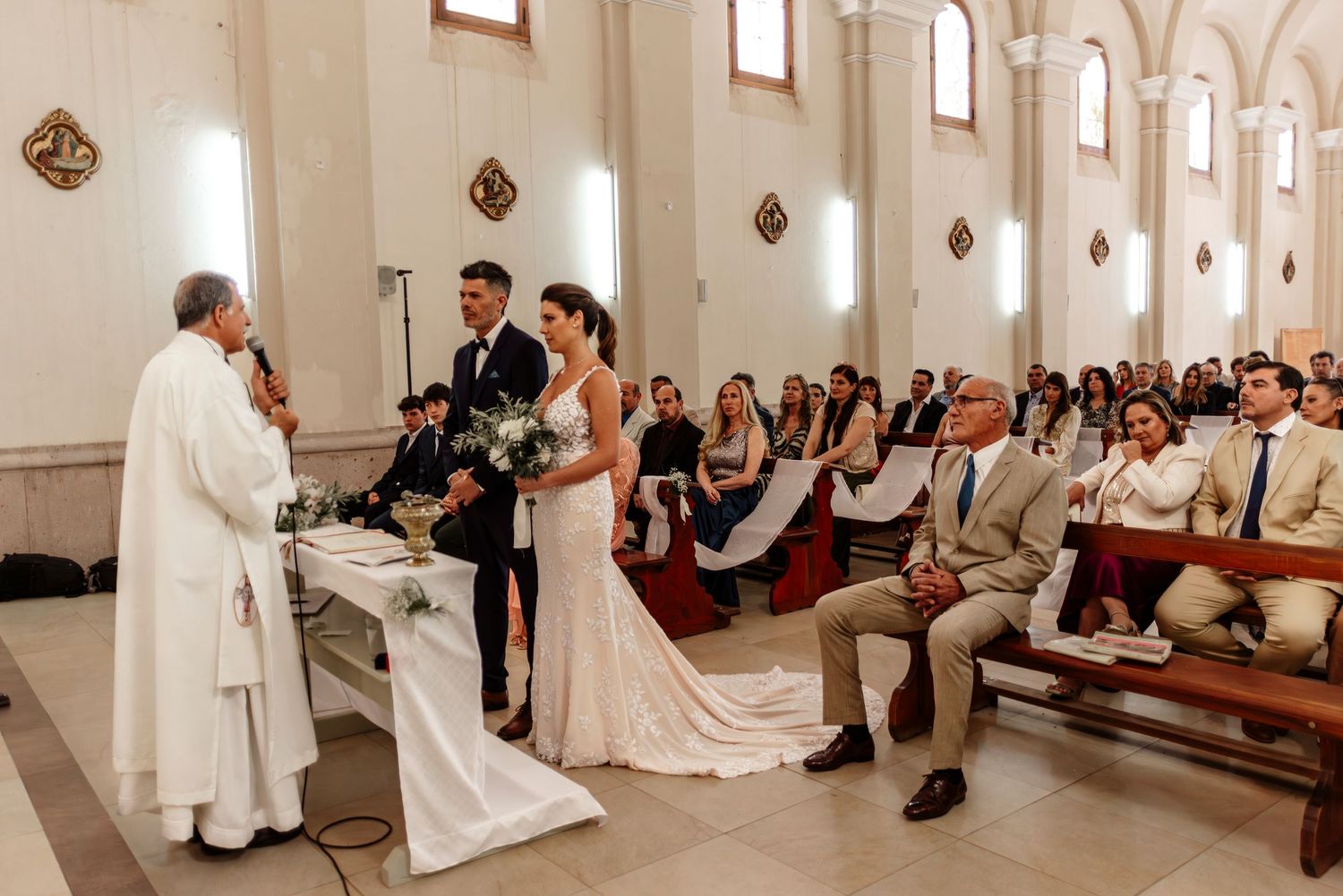 A panoramic view of a Catholic wedding ceremony inside a bright white church with wooden pews and arched windows.