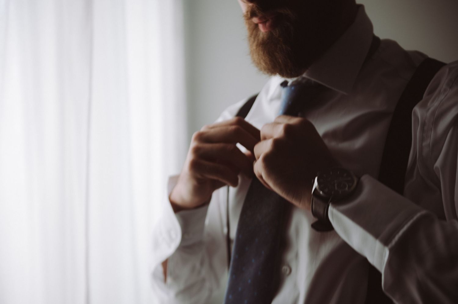 Close-up view of someone adjusting their tie while wearing a white dress shirt in moody lighting near a window.