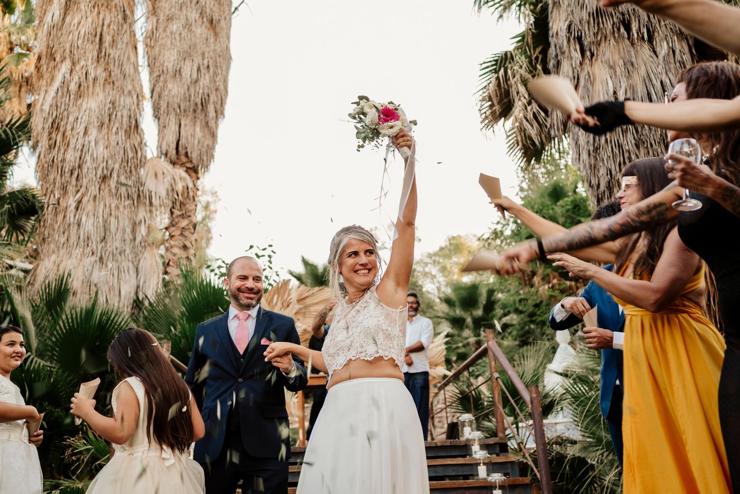 Joyful outdoor wedding celebration under palm trees as couple celebrates with confetti and flowers in the air.