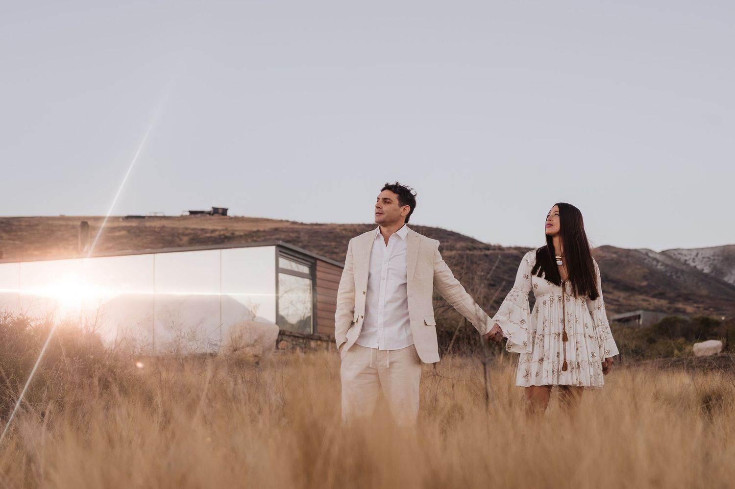 Pareja tomada de la mano en un campo dorado al atardecer, cerca de un moderno edificio de cristal con las montañas de fondo. preboda mendoza