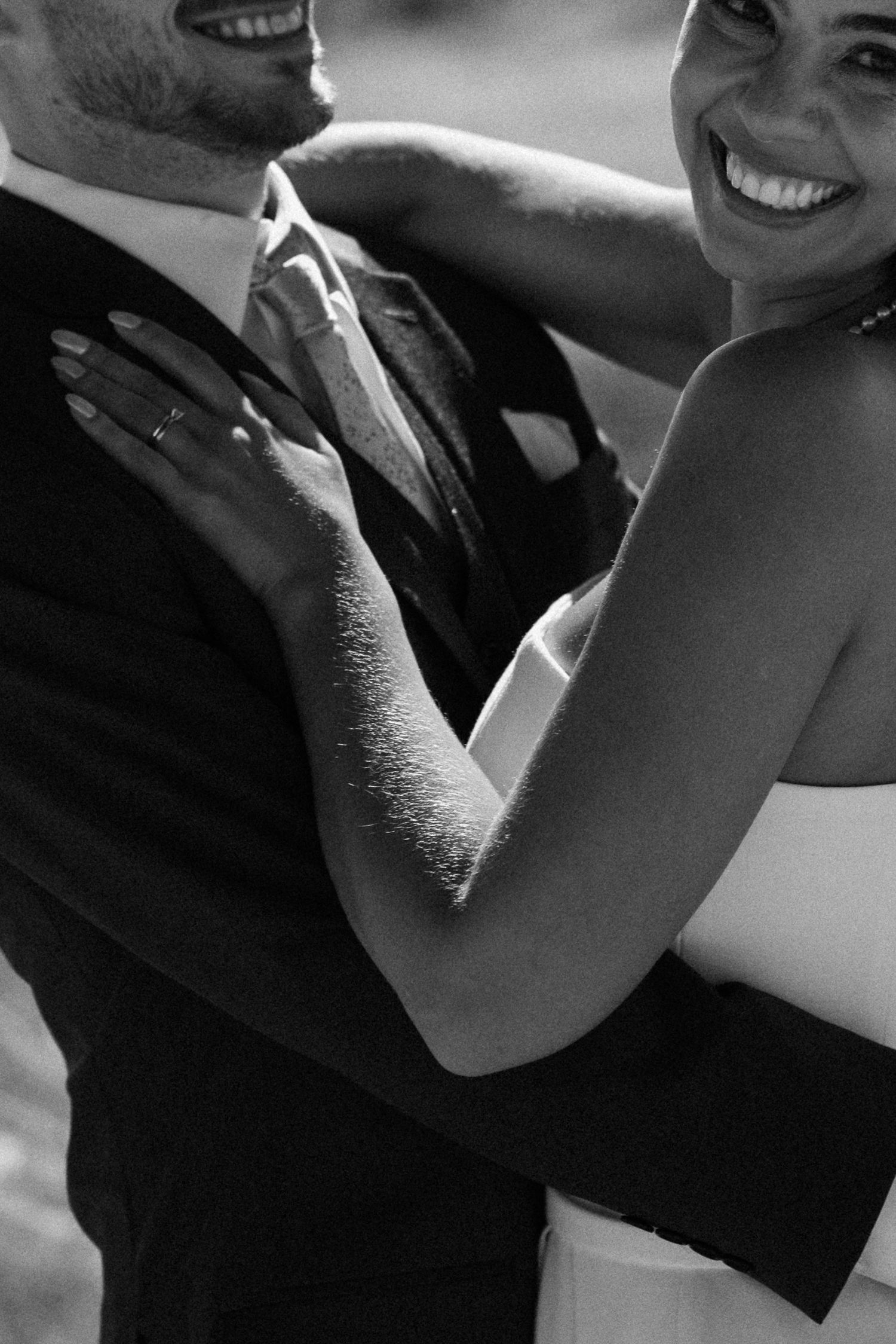 Black and white photo of a smiling bride and groom embracing, showing wedding rings, in elegant formal attire.