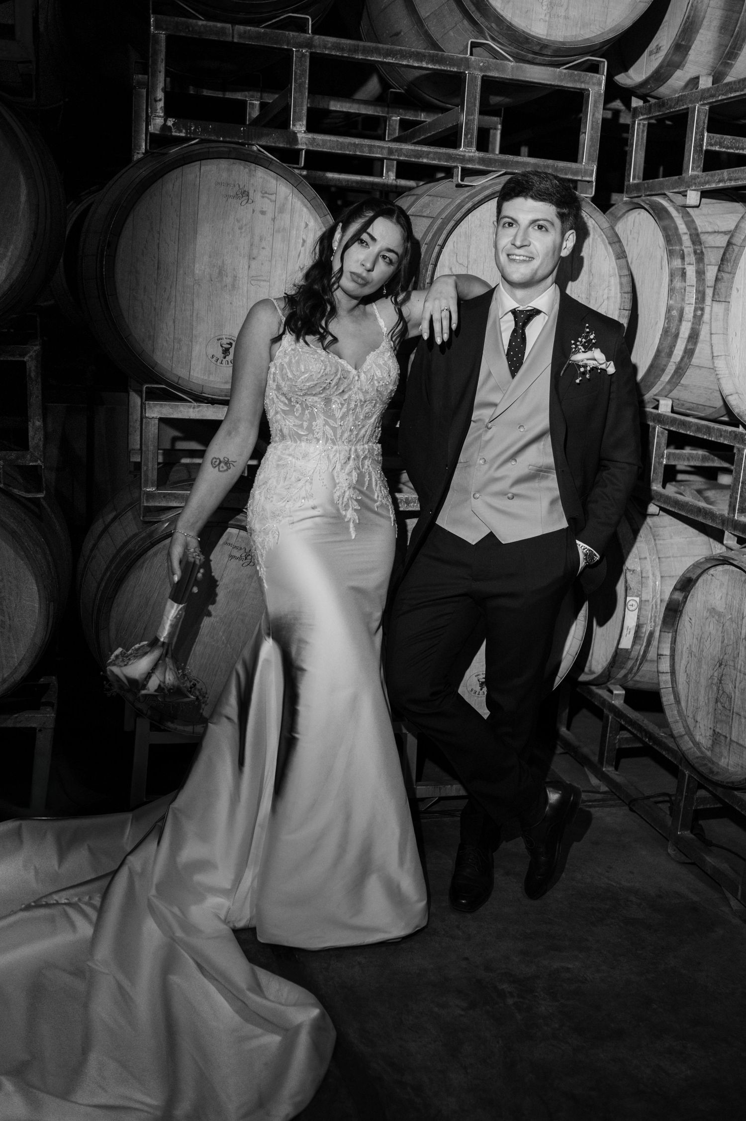 Bride and groom pose elegantly among wine barrels in a winery in this stunning black and white wedding portrait.