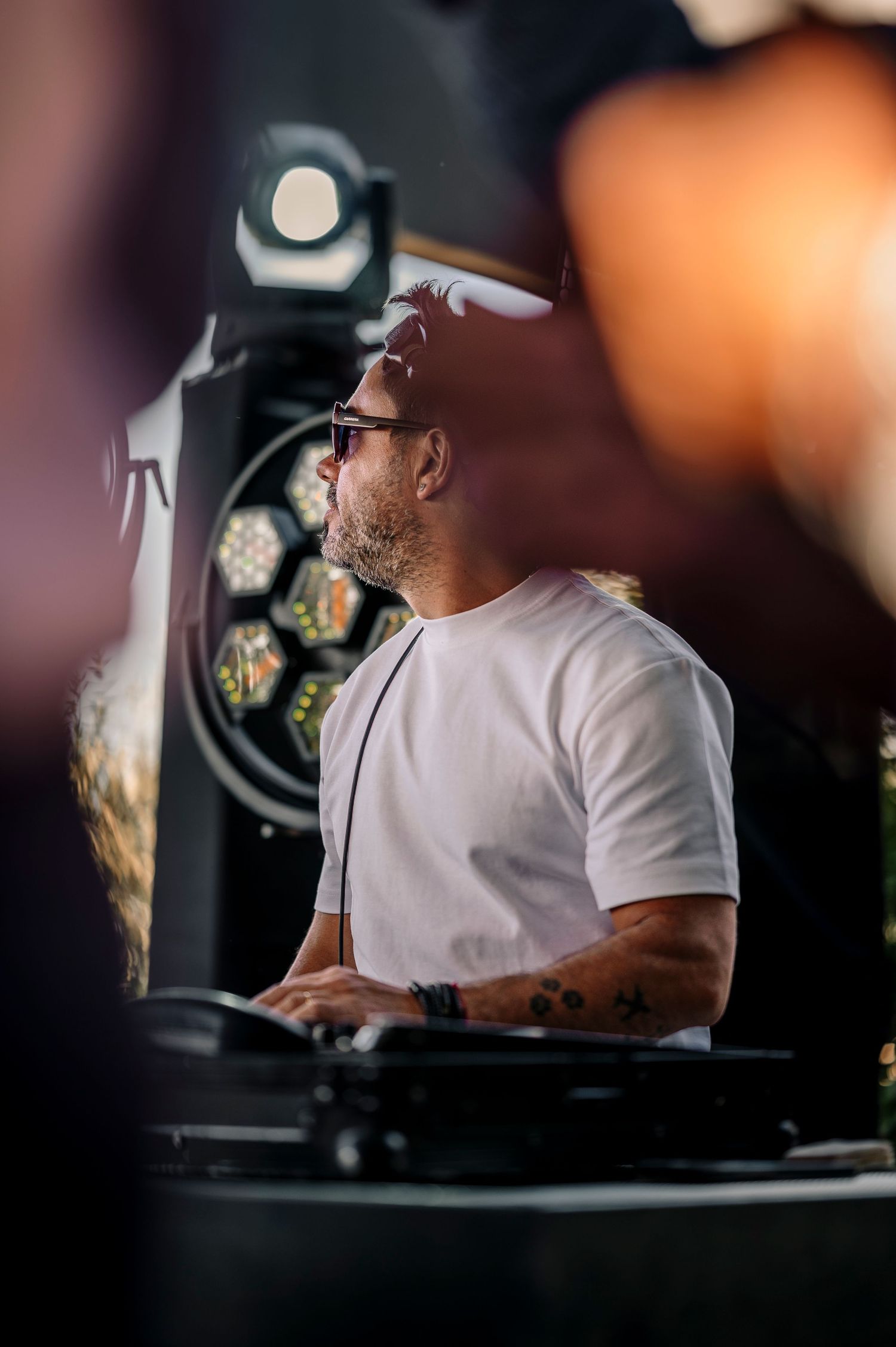 A person in a white t-shirt and cap sits at a dimly lit control panel or desk in a dark room.