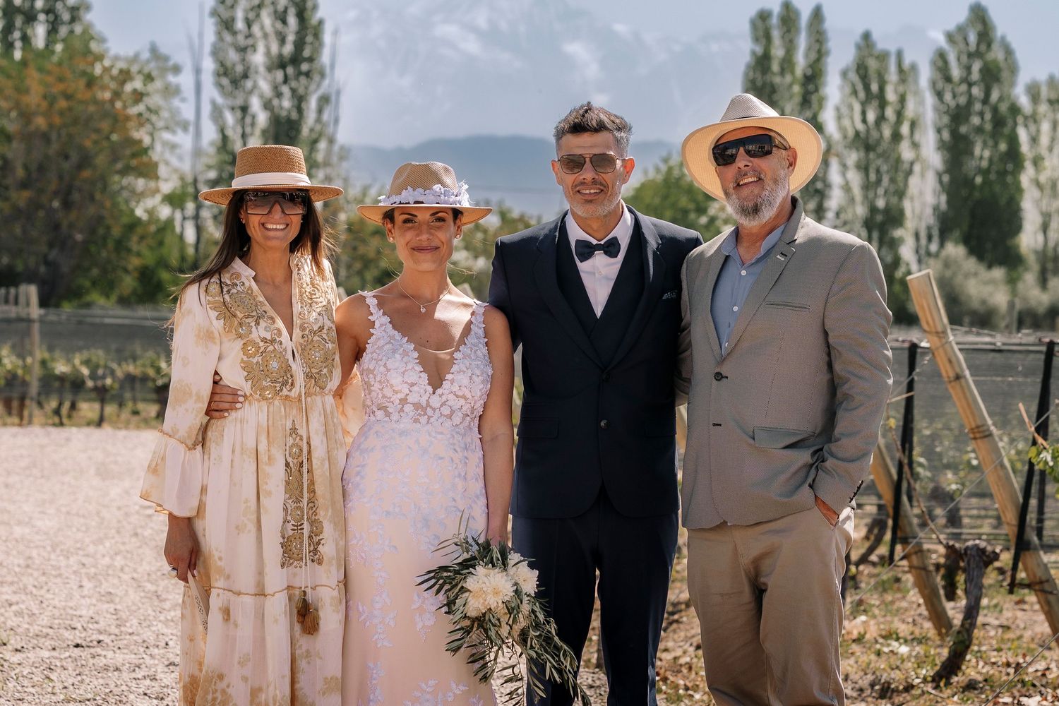 A group of people in formal attire pose together outdoors at a rustic venue on a sunny day.