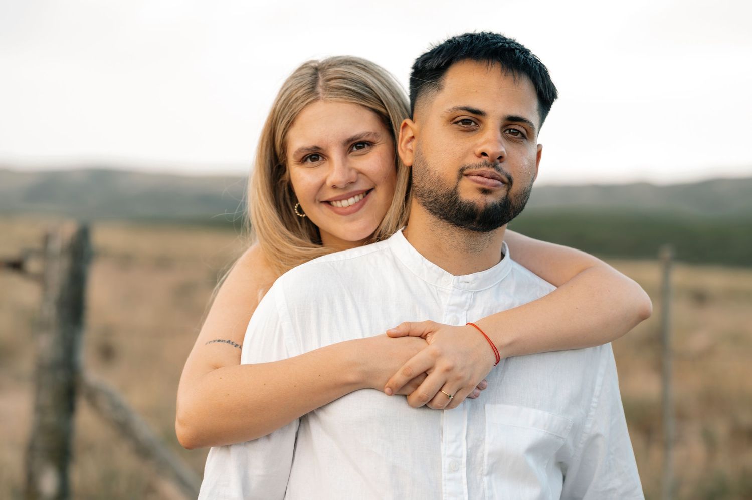 A couple in white outfits embraces lovingly in a scenic outdoor setting with rolling hills in the background.