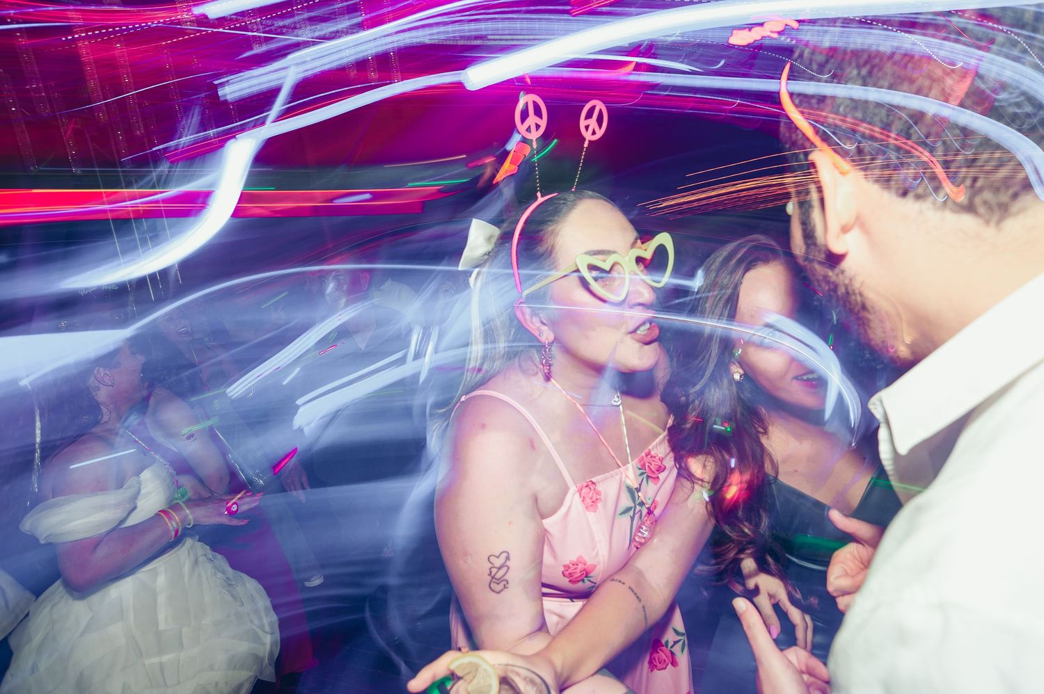 A wedding couple shares a romantic moment on a dance floor illuminated by vibrant pink and purple lights.