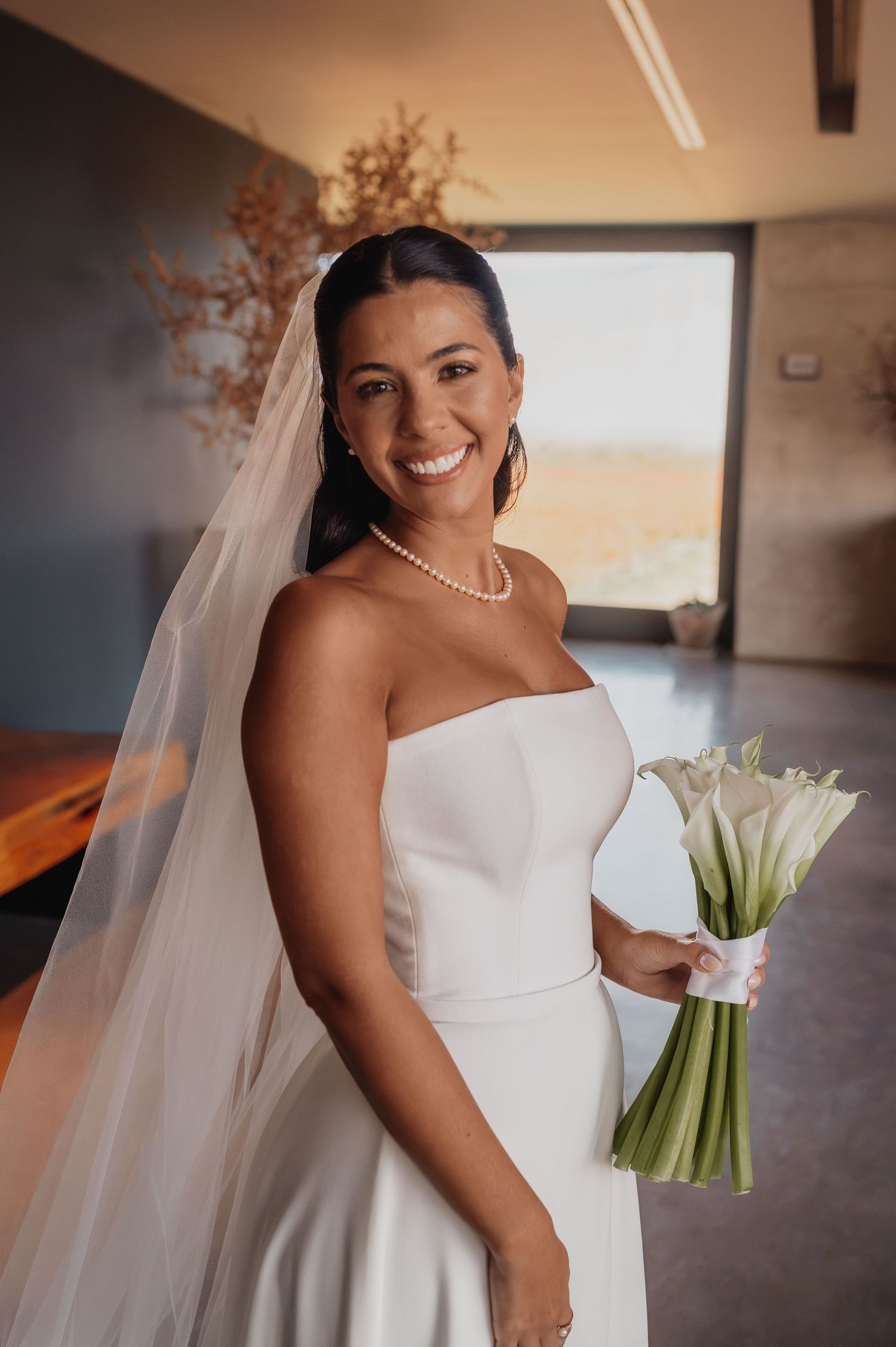Smiling bride in strapless white gown, pearl necklace, veil, holding white calla lily bouquet indoors. fotógrafos de boda Mendoza
