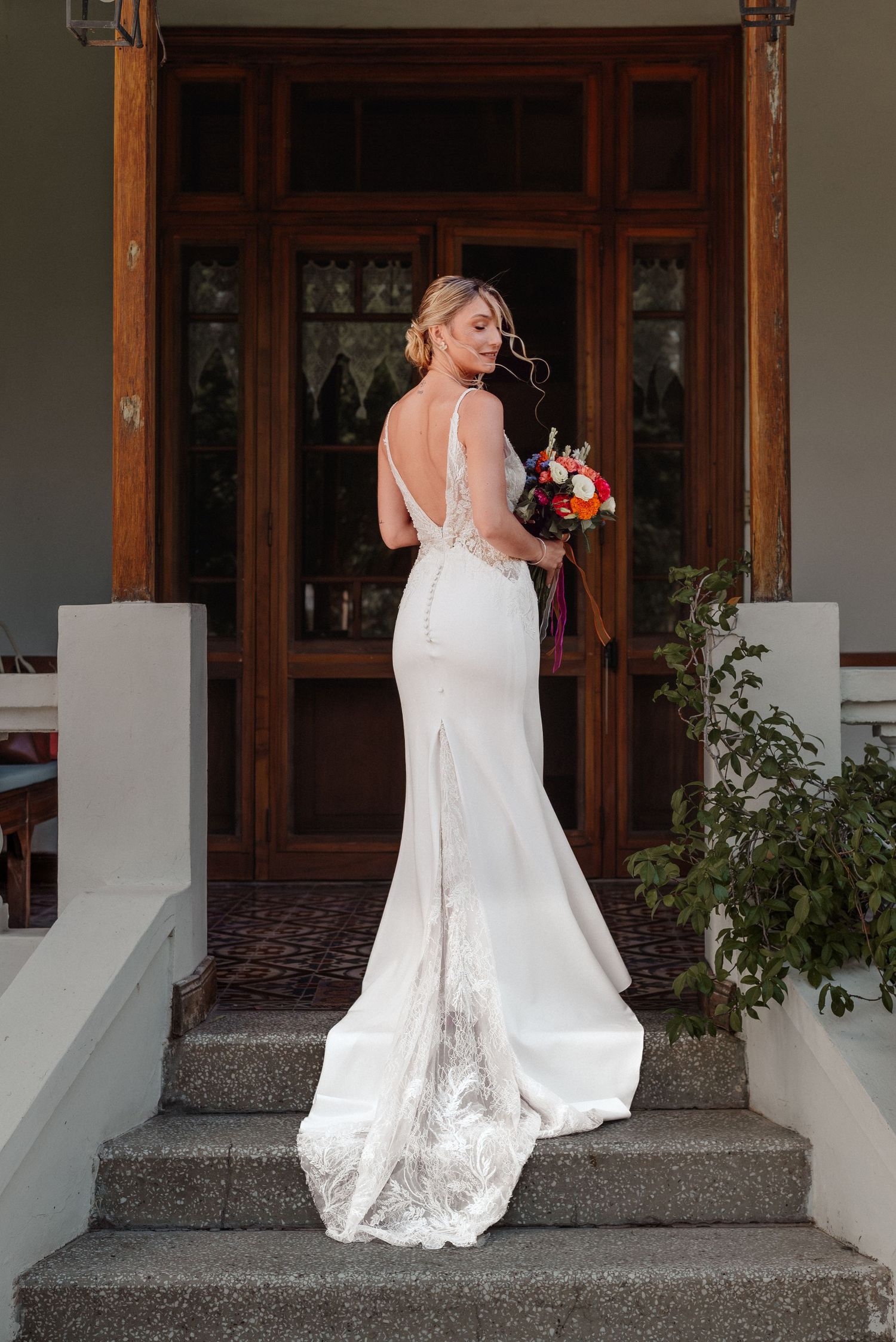 Bride in elegant backless white gown with lace train holds colorful bouquet on steps of a grand entrance.