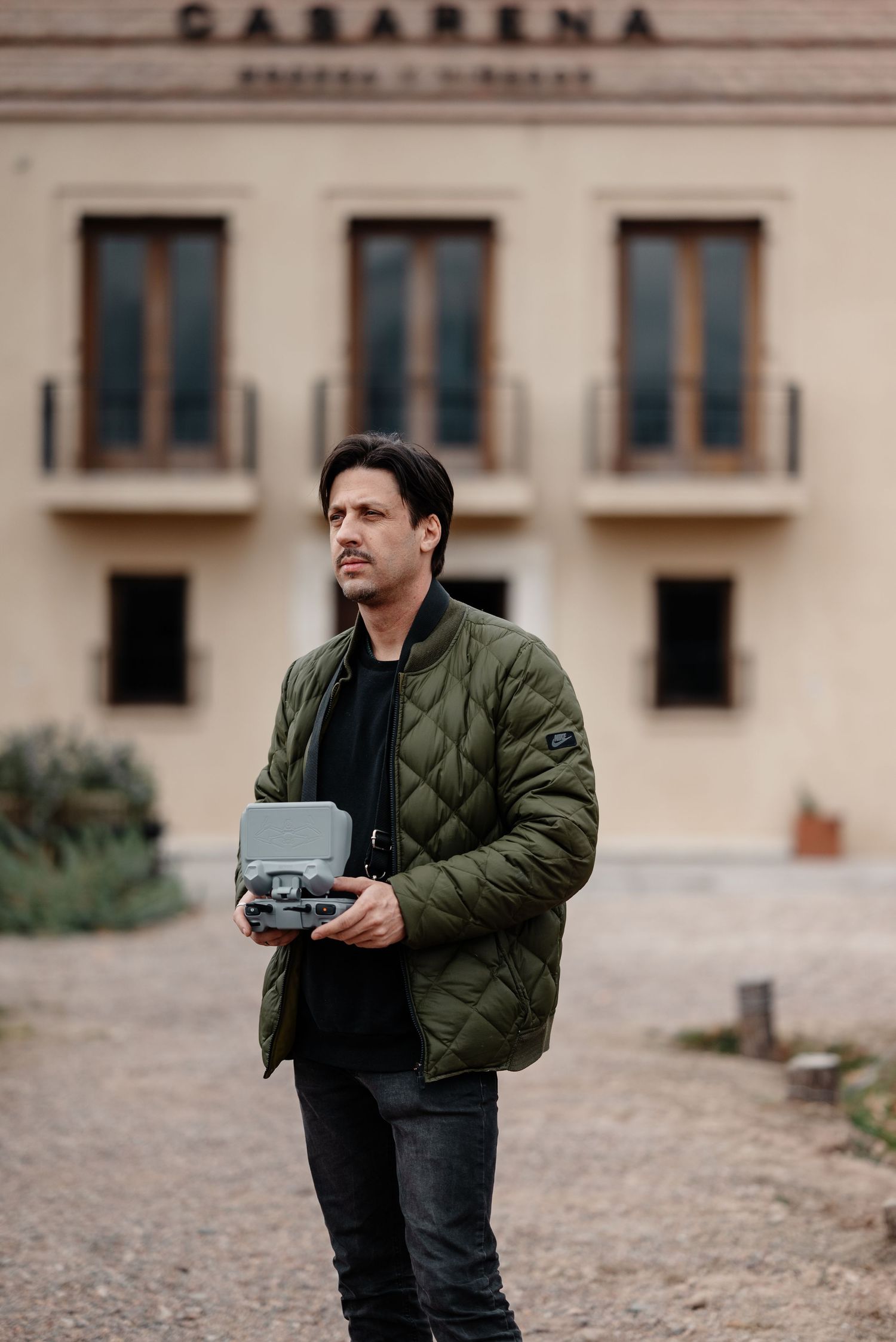 Person wearing green quilted jacket holds drone controller device while standing outside building.