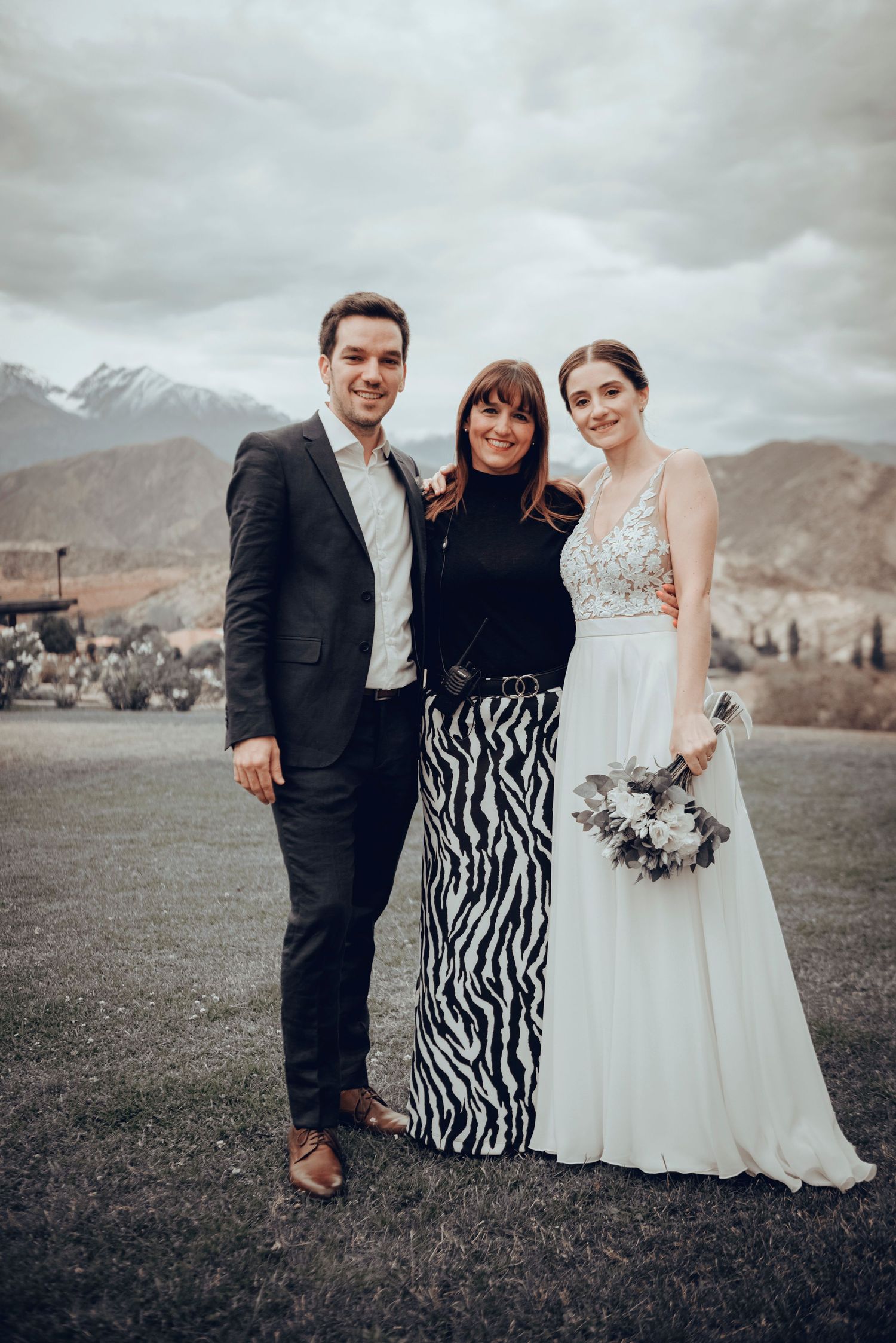 A wedding group poses together on a grassy hillside with mountains in the background as clouds roll in overhead.