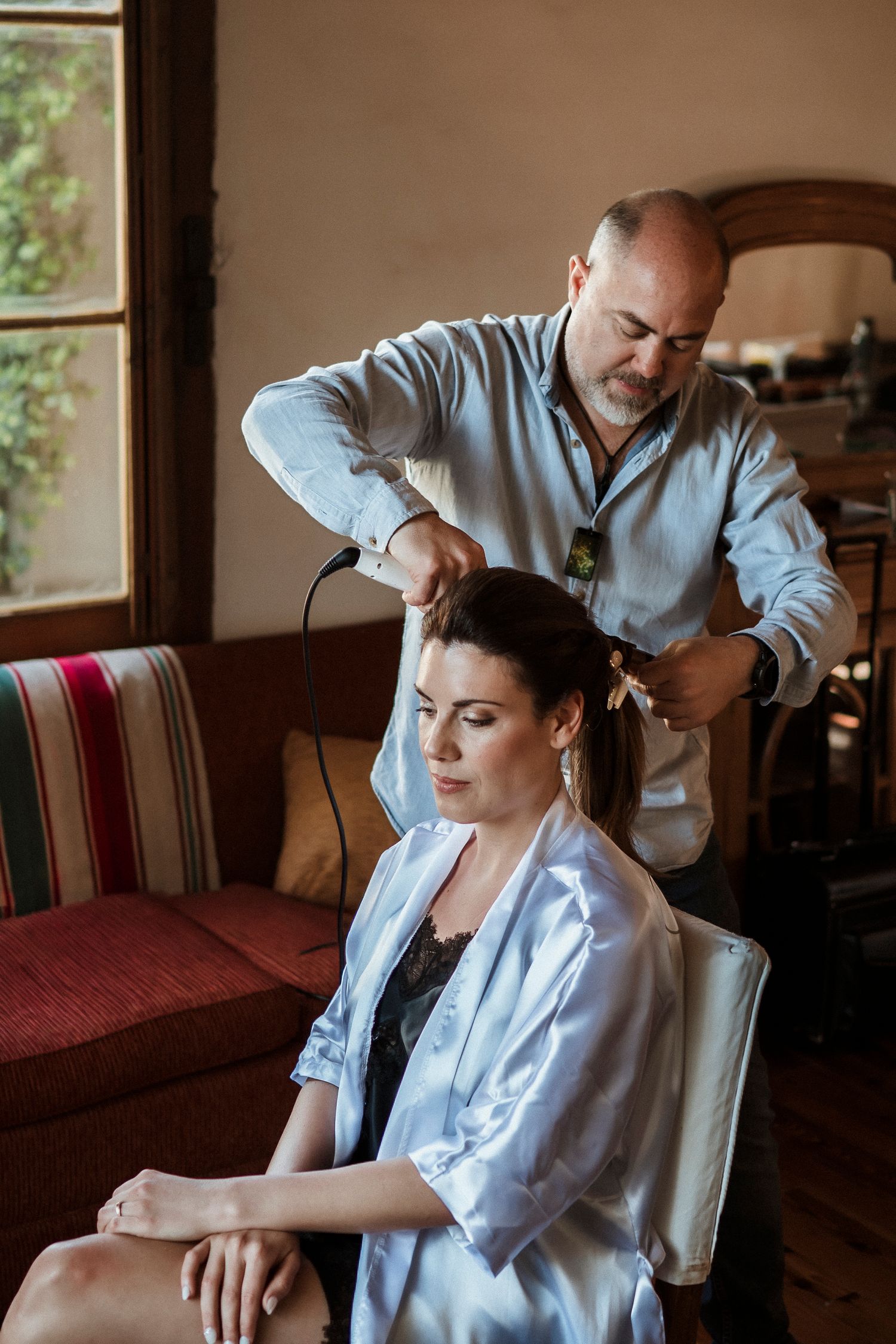 A hairstylist works on styling a client's hair in a robe while seated on a red striped couch near a window.