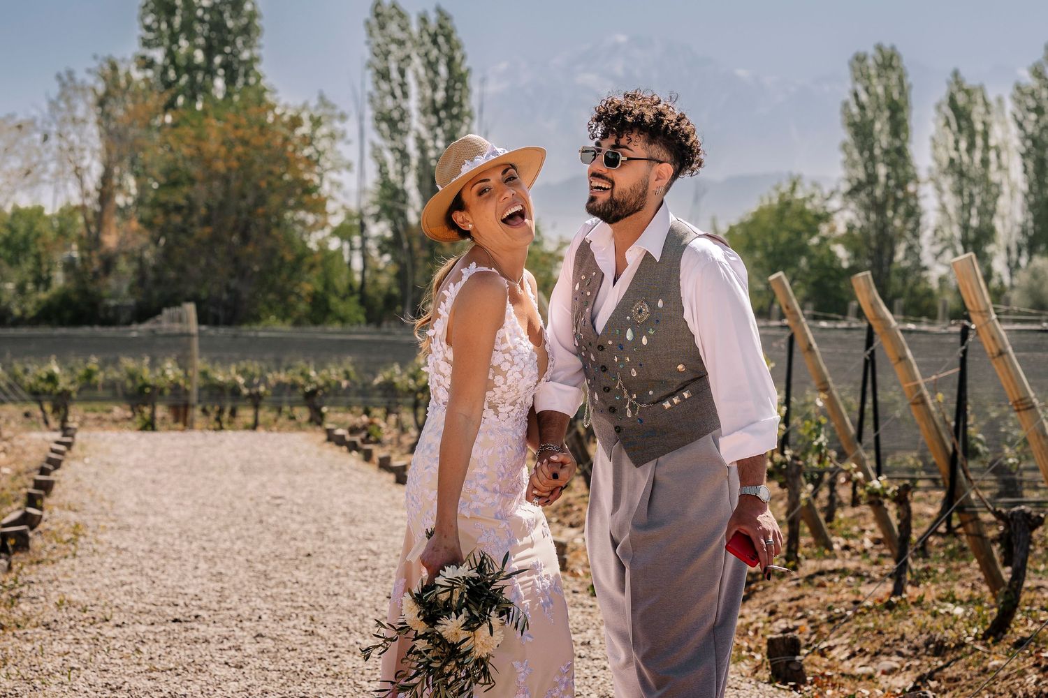 A couple laughs together while walking through rows of grapevines in a rustic vineyard setting.
