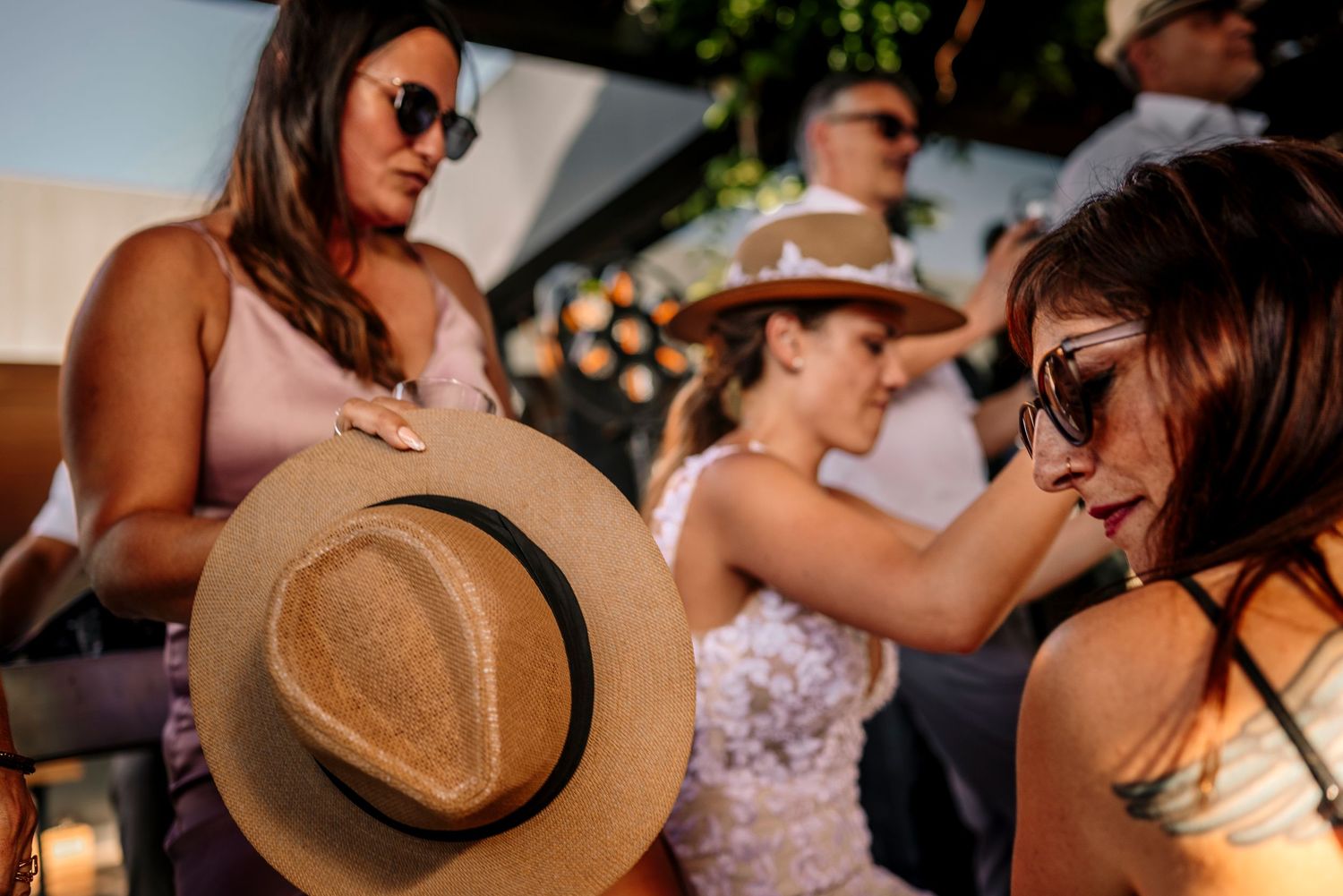 A group of wedding guests dance and celebrate at an outdoor reception while wearing straw hats and summer attire.