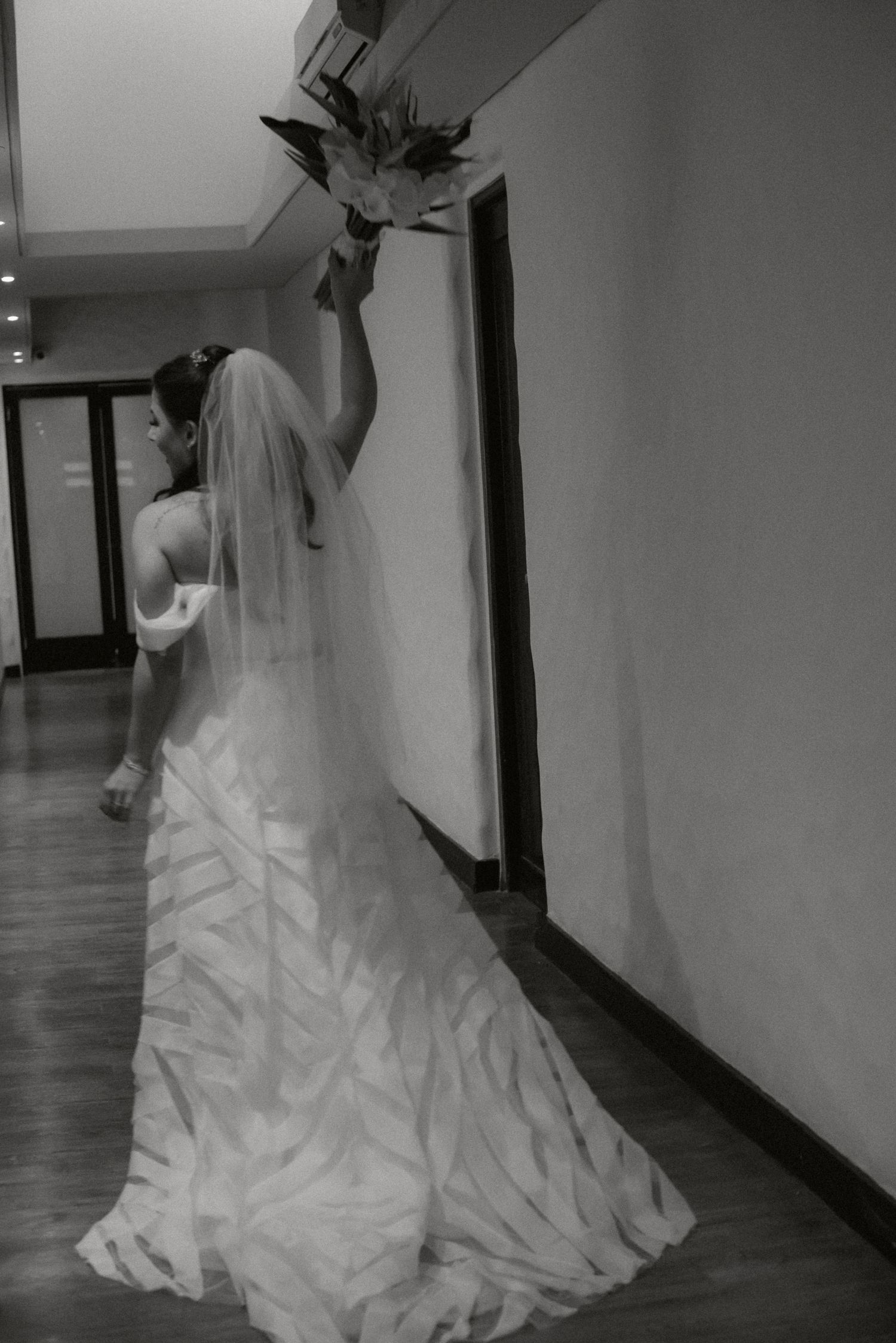 A bride in a flowing white wedding gown and veil reaches up to touch a light fixture in a hallway.