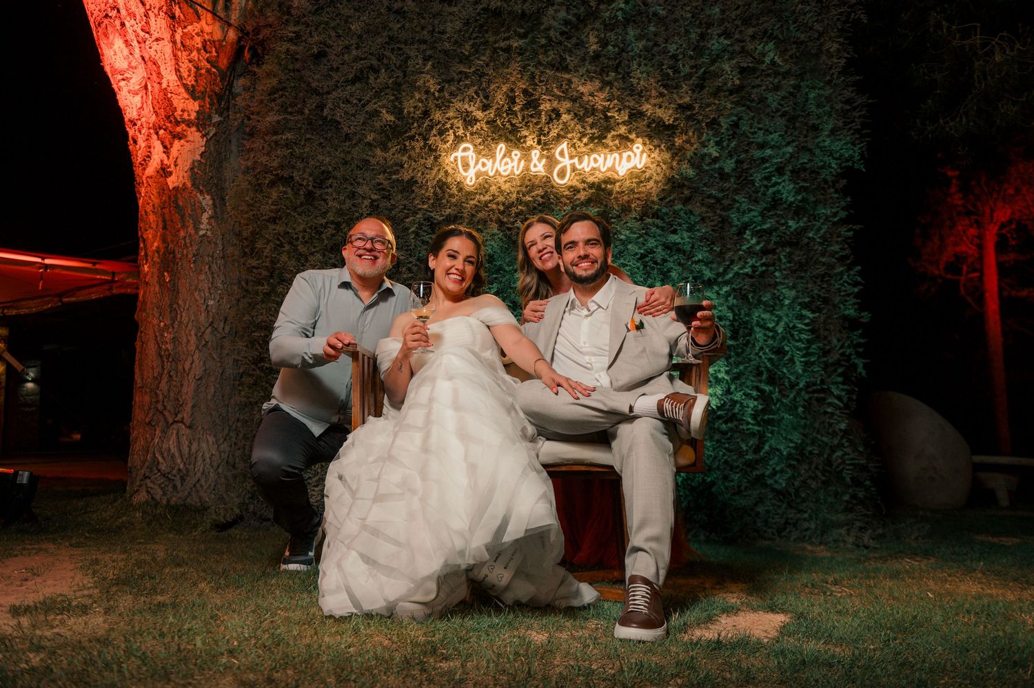 A wedding party sits on the grass during an outdoor evening celebration with warm lighting and trees in the background.