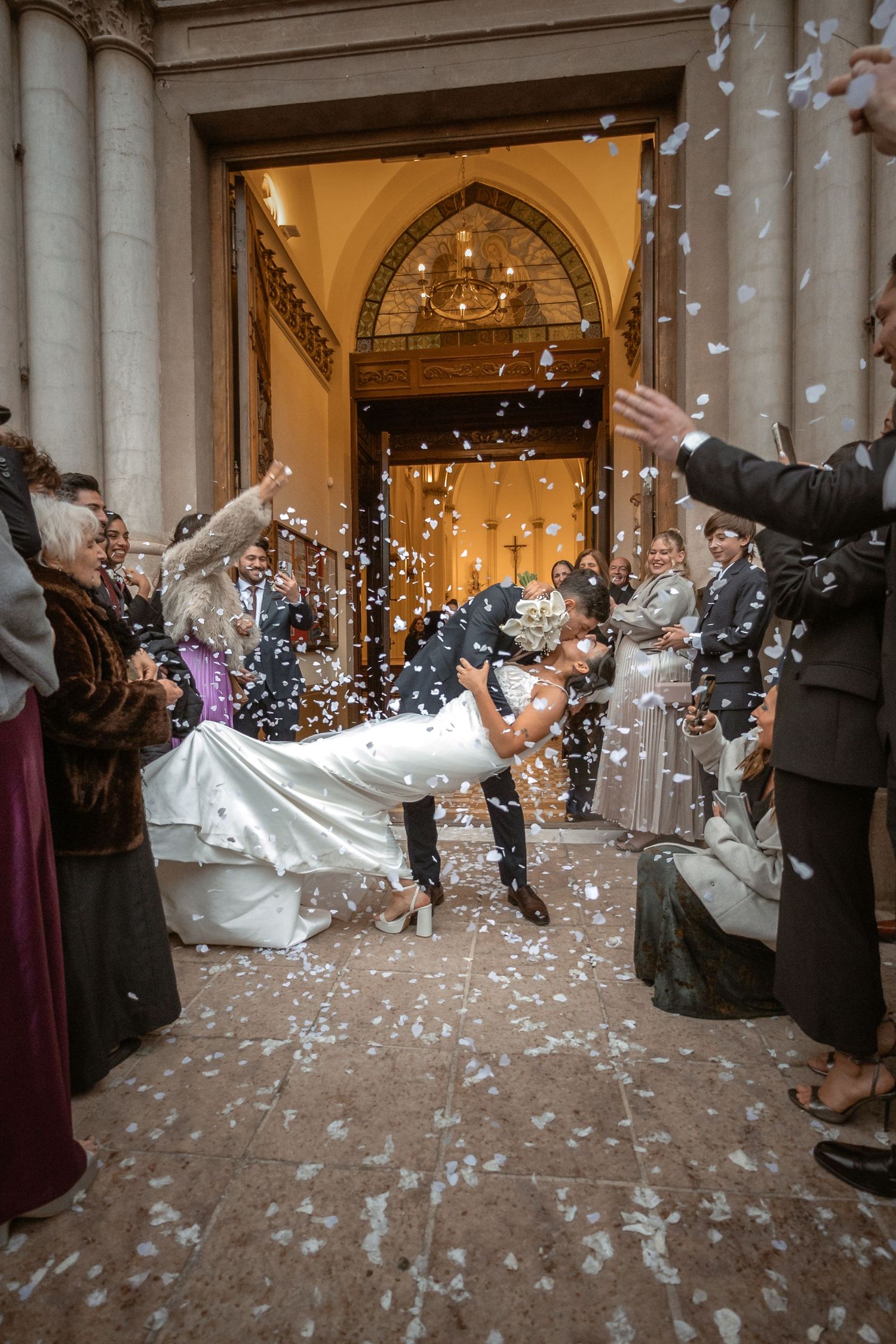 Groom dips bride outside church entrance as wedding guests toss white confetti in joyful celebration.