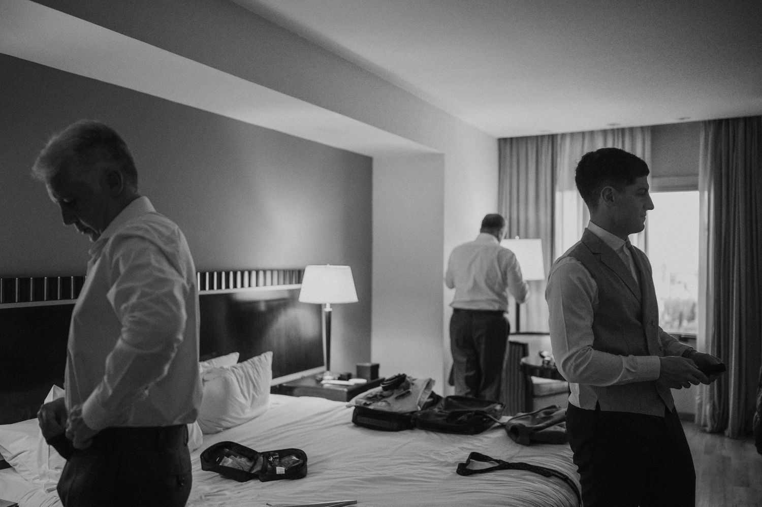 Black and white photo of groomsmen getting ready in a hotel room before a wedding ceremony.