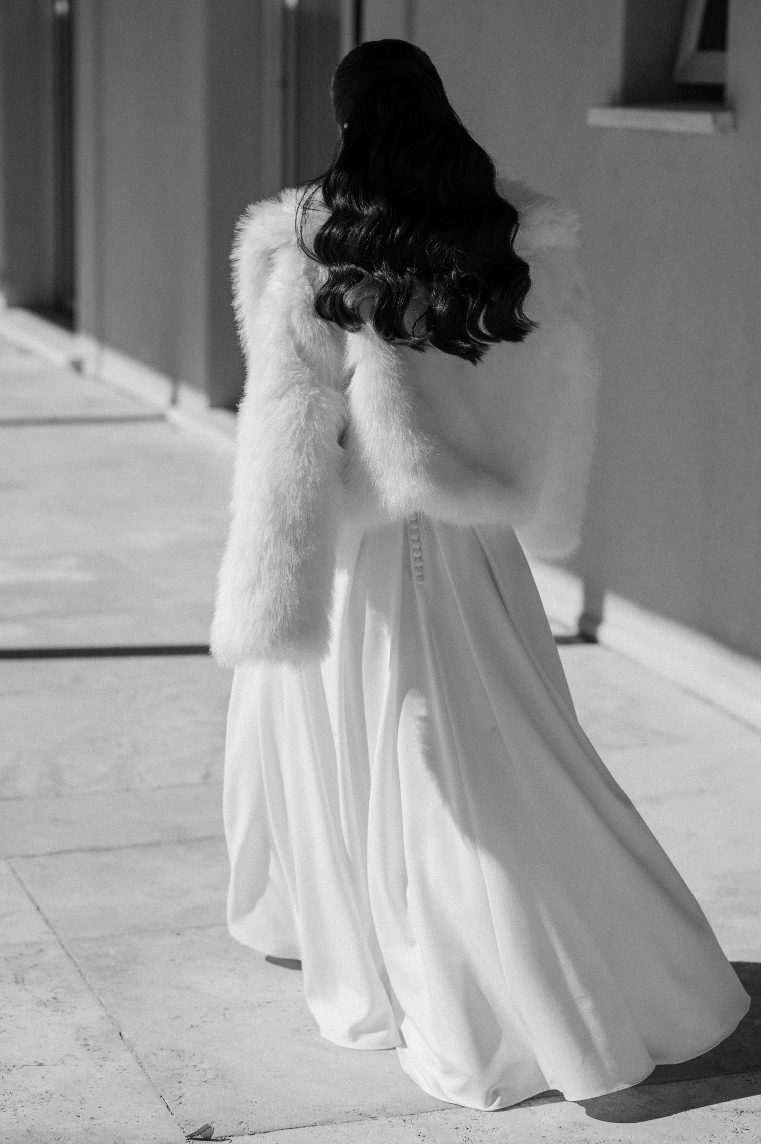 Black and white photo of a bride in a white gown and fur stole, viewed from behind, with long dark curly hair.