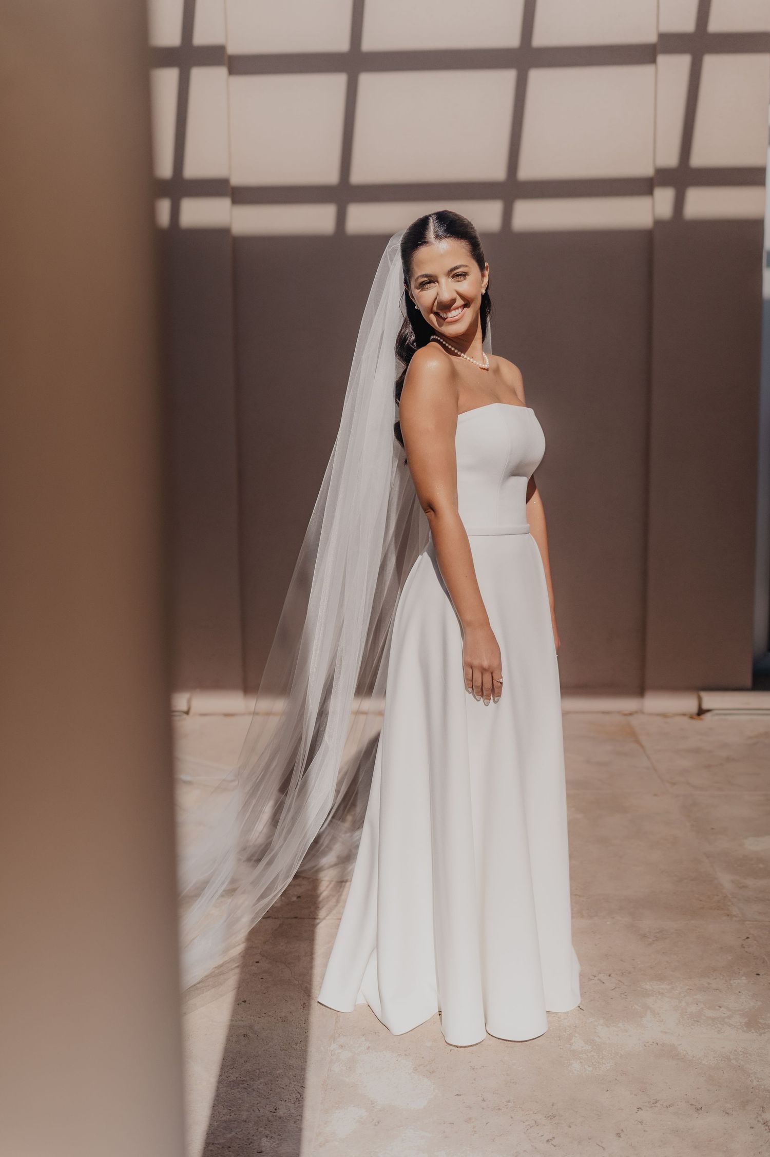Smiling bride in a strapless white gown with a long flowing veil poses elegantly in a sunlit modern venue.