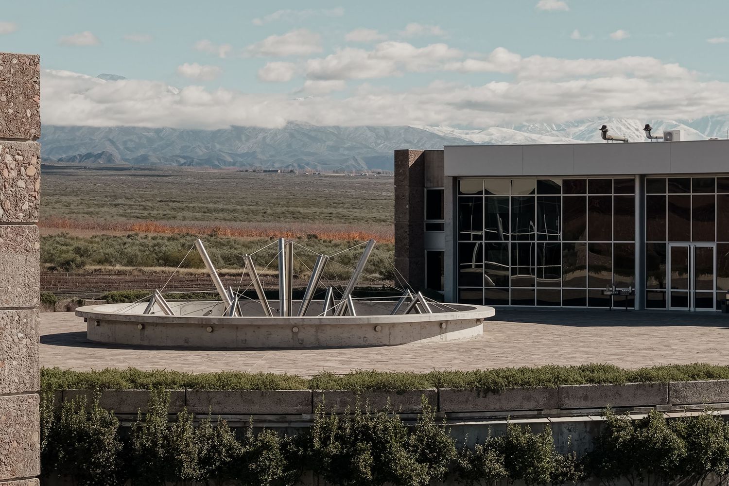 Modern building with circular fountain plaza overlooking vast desert landscape and snow-capped mountains under cloudy sky.