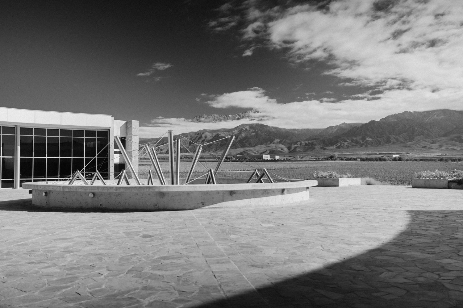 Black and white photo of a modern building with geometric steel sculptures, mountain backdrop, and dramatic cloudy sky.