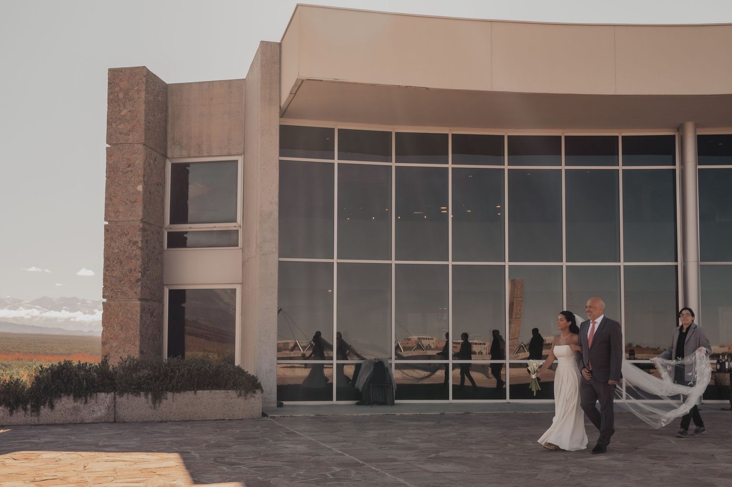 Bride and groom walk hand-in-hand outside a modern glass building venue during golden hour wedding celebration.