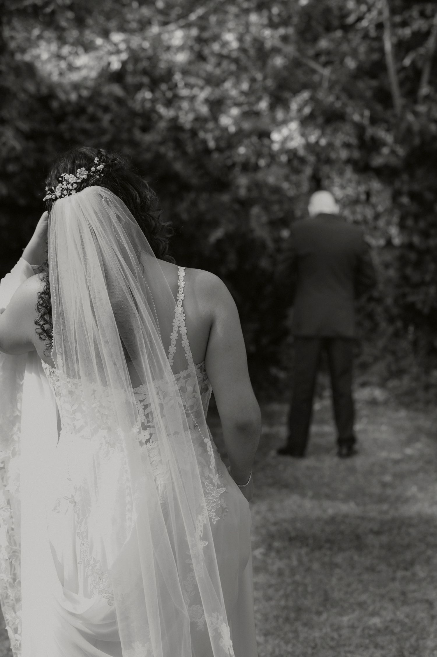 Black and white wedding photo showing bride in lace veil facing groom from behind in outdoor garden setting.