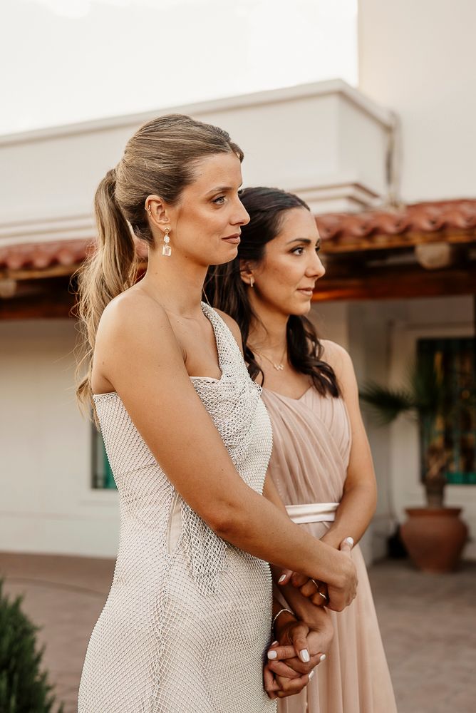 Two women in formal dresses stand together at an outdoor venue with white walls and terracotta roof tiles. wedding in mendoza
