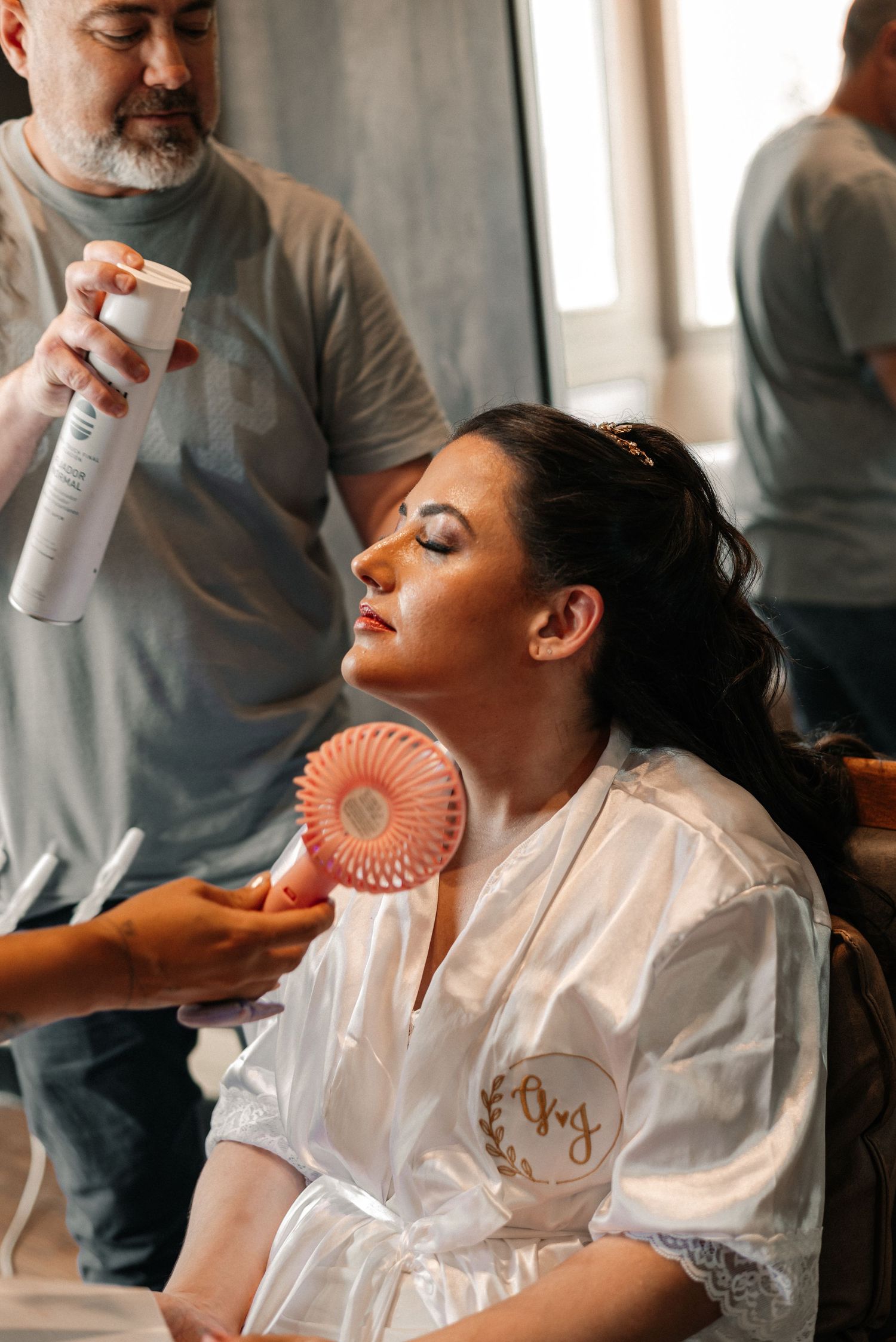 A person in a white satin robe gets their hair styled while holding a small pink handheld fan.