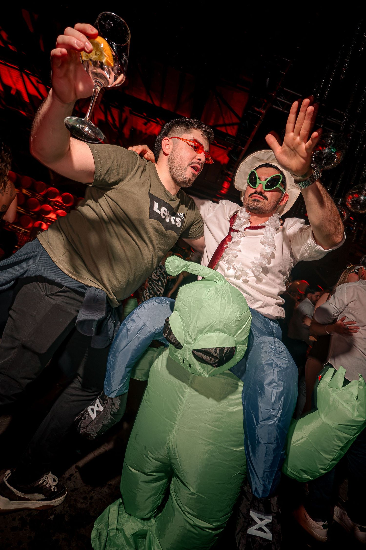Two men in alien-themed costumes celebrate at a nightclub party, one holding a cocktail glass aloft.