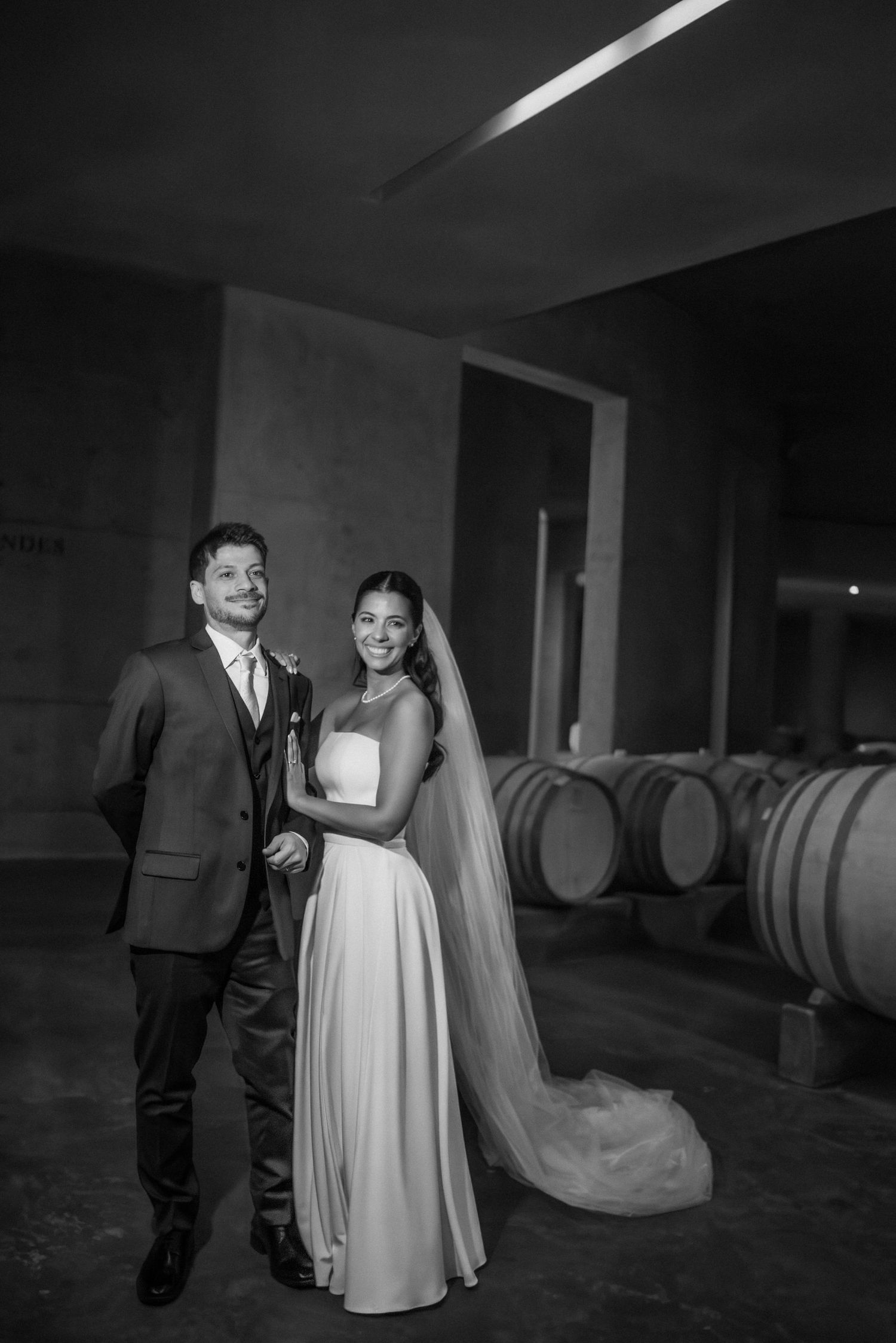Black and white wedding photo of bride in veil and groom in suit posing in a winery with oak barrels. fotógrafos de boda Mendoza