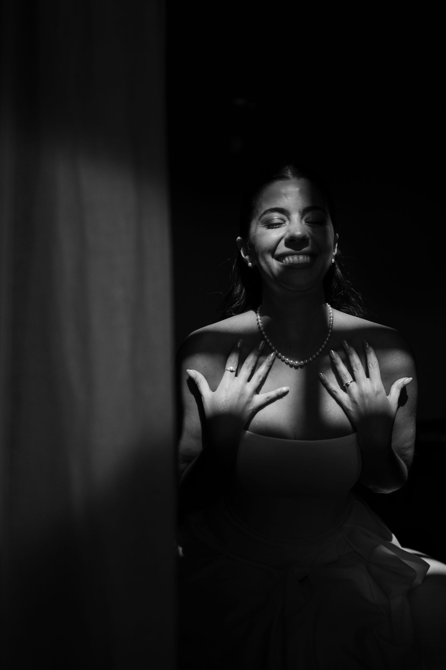 Black and white photo of a smiling woman in a strapless top and pearl necklace, hands on chest, eyes closed. fotógrafos de boda Mendoza