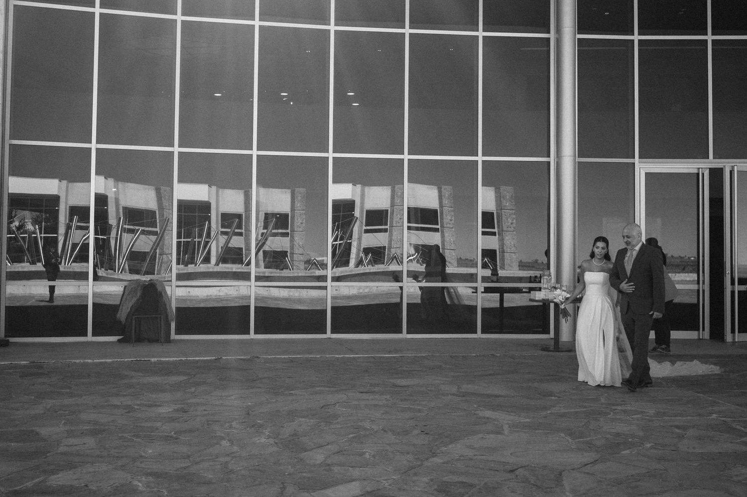 Black and white photo of a bride and groom standing outside a large glass building at their wedding reception.