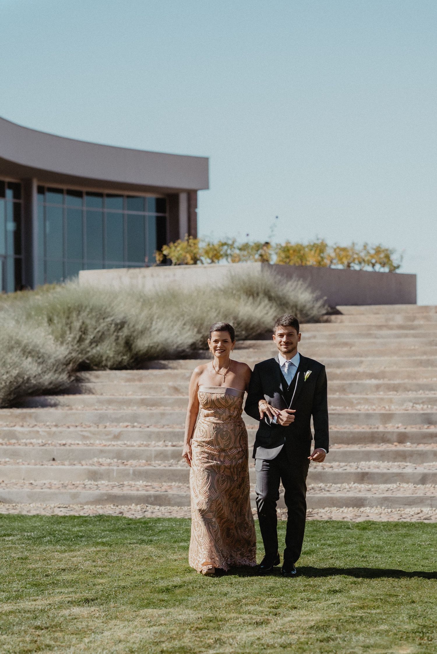 Couple in formal attire walking on green lawn outside modern venue with stone steps on sunny day.
