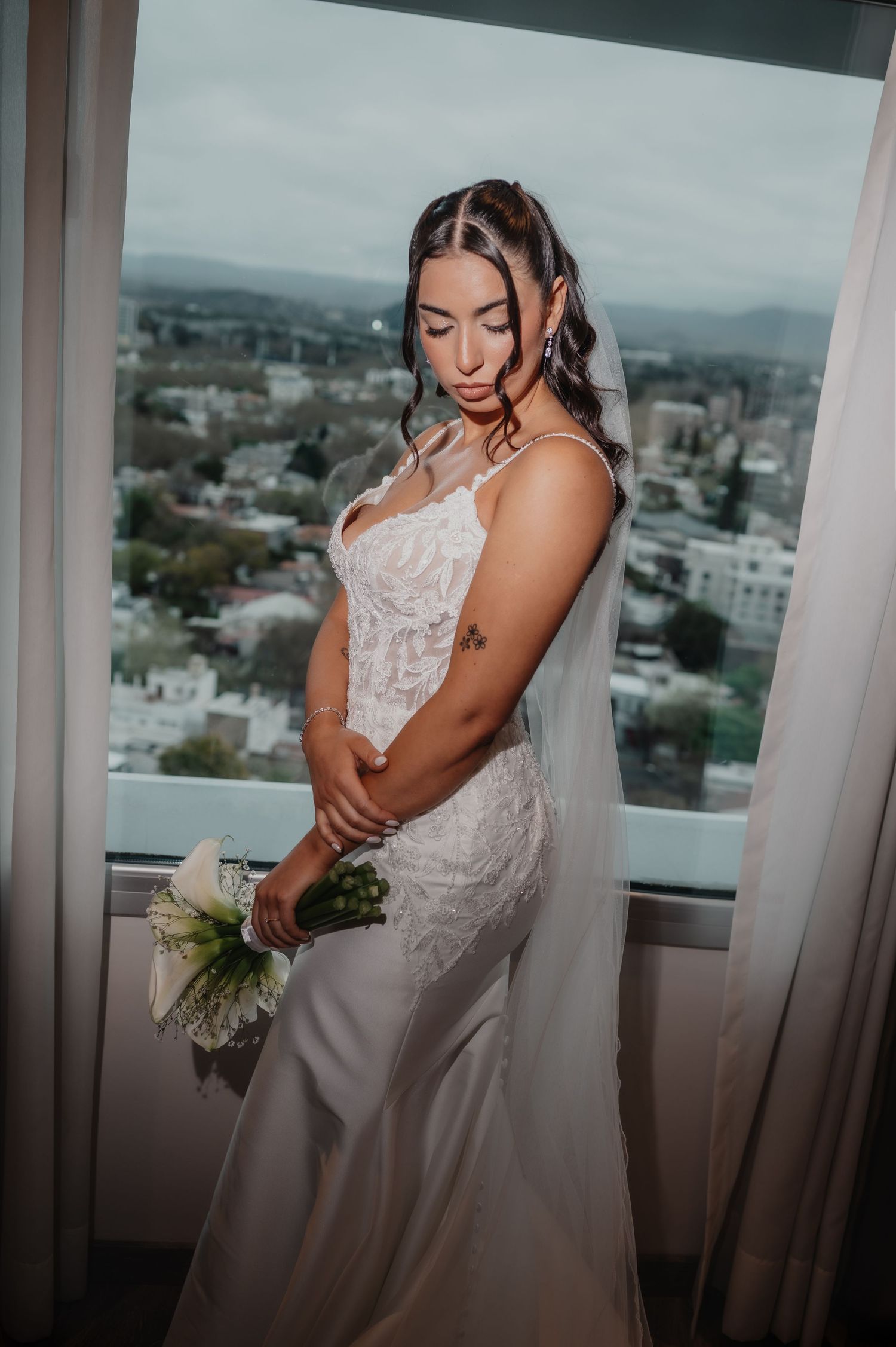 a novia, con un vestido de sirena de encaje blanco, sostiene un ramo junto a la ventana de un rascacielos con vistas a la ciudad de mendoza