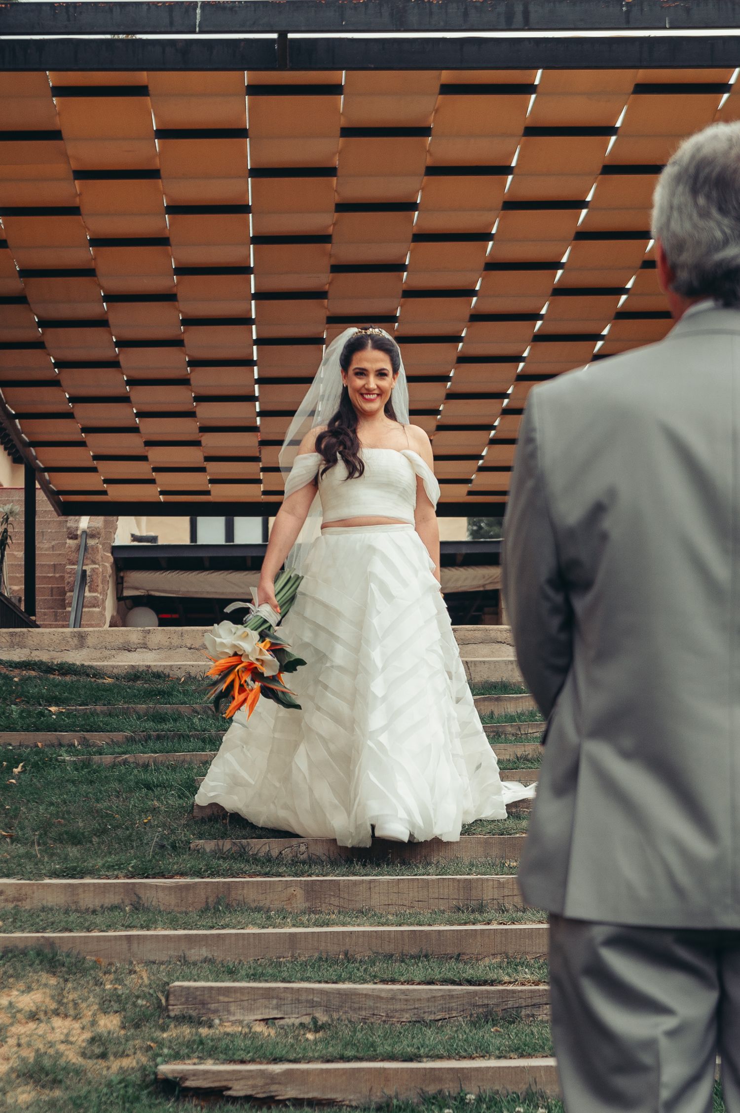 A bride in a flowing white dress walks up tiered outdoor steps while a person in a gray suit watches from behind.