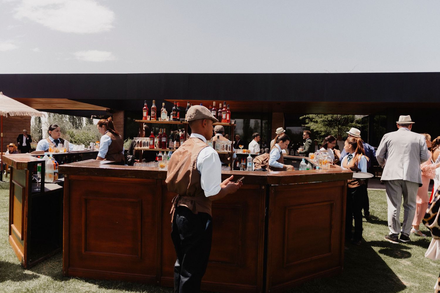 A rustic outdoor wedding bar setup with wooden counters and bartenders serving drinks to guests at a garden reception.